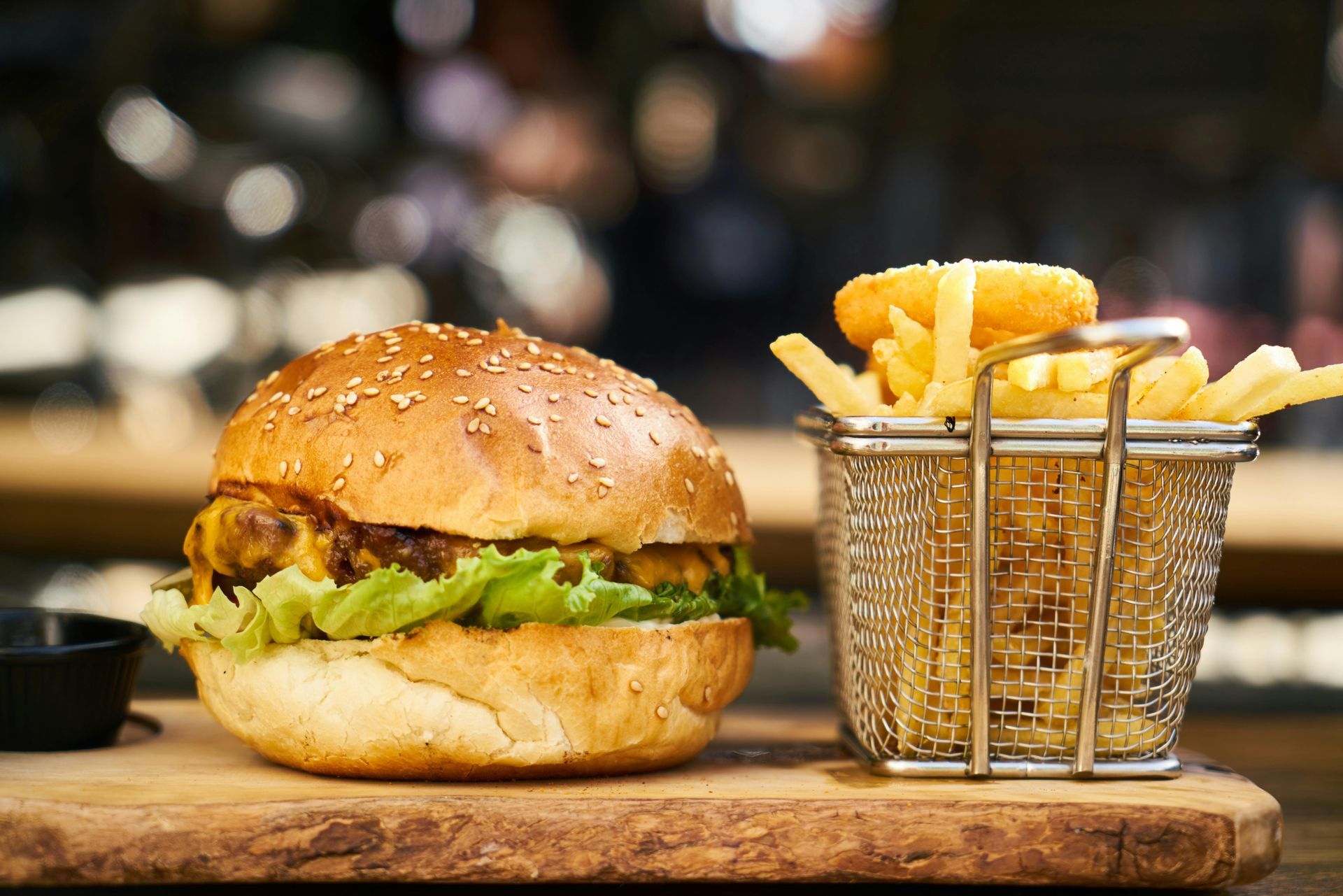 A sesame seed bun cheeseburger with lettuce next to a metal wire basket of french fries on a wooden serving board.