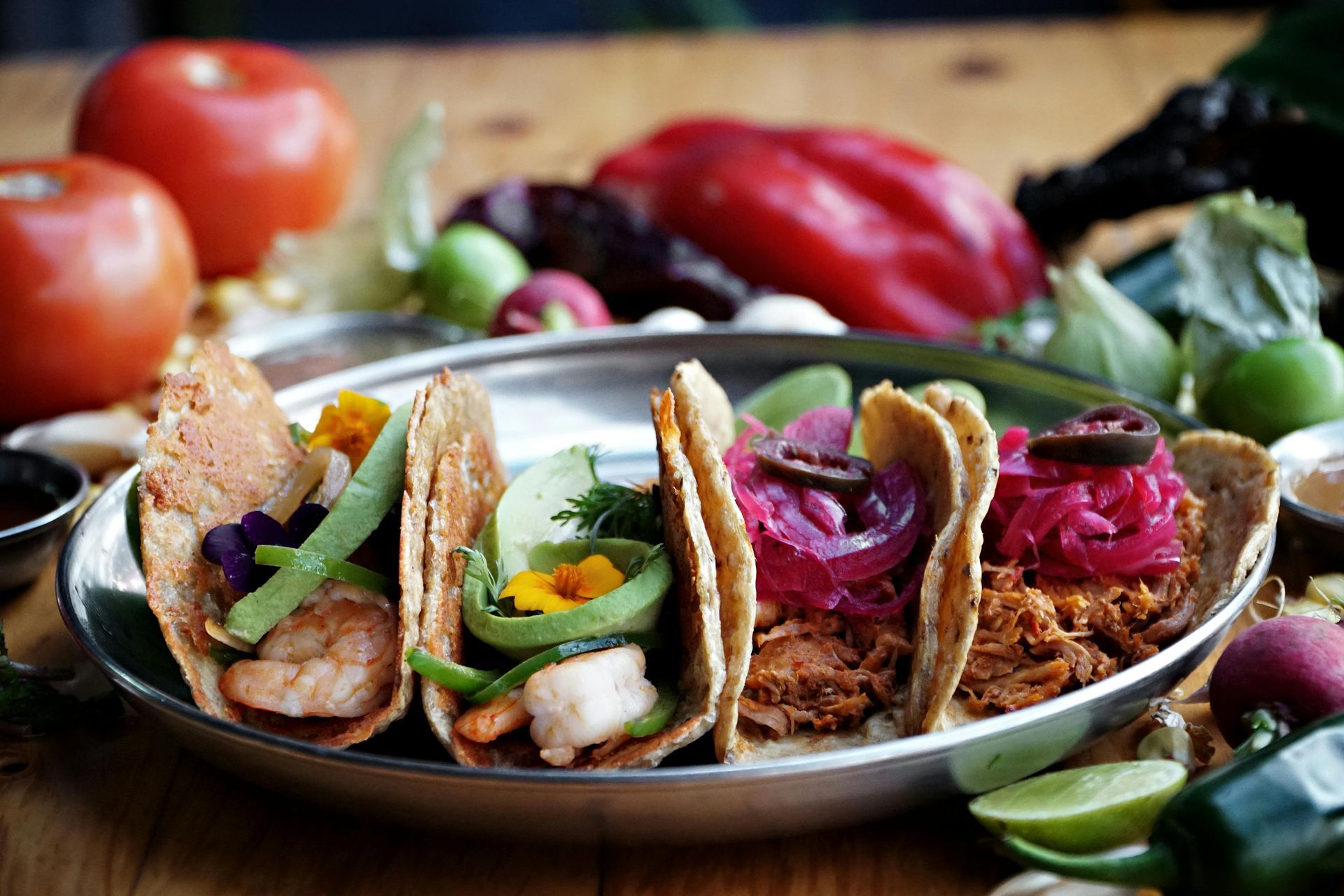 Tacos on a metal plate, assorted fillings, surrounded by vegetables like tomatoes, peppers, and limes.