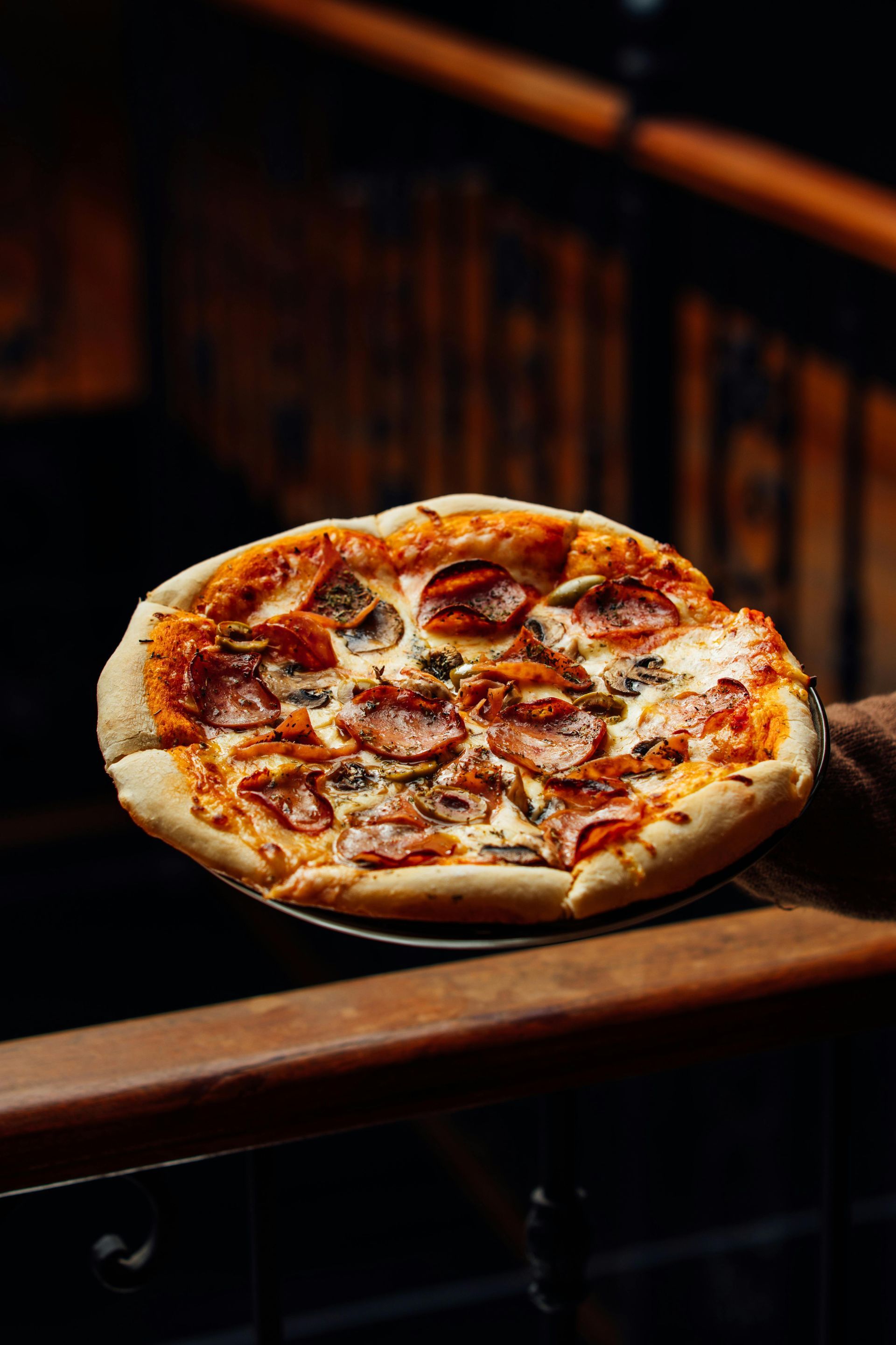 Pepperoni pizza on a metal tray, held by a person on a wooden handrail, dimly lit.