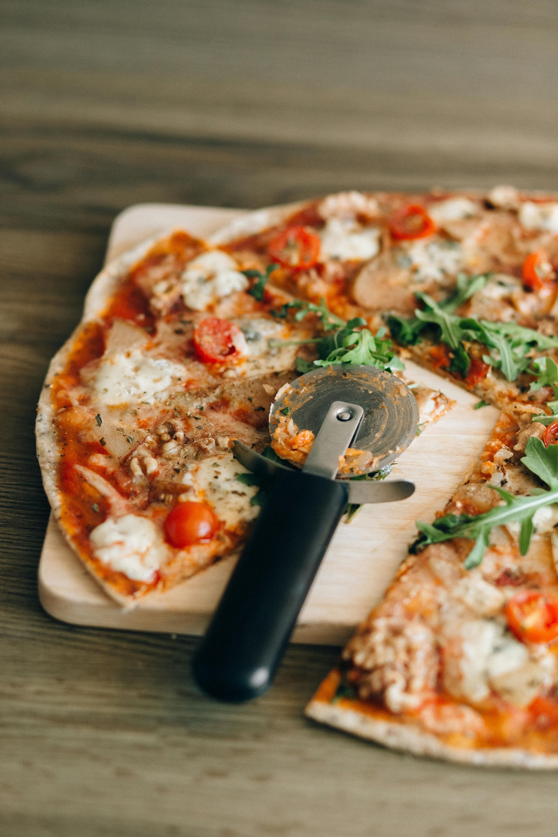 A pizza with cherry tomatoes and greens on a wooden board, with a black-handled pizza cutter resting on top.