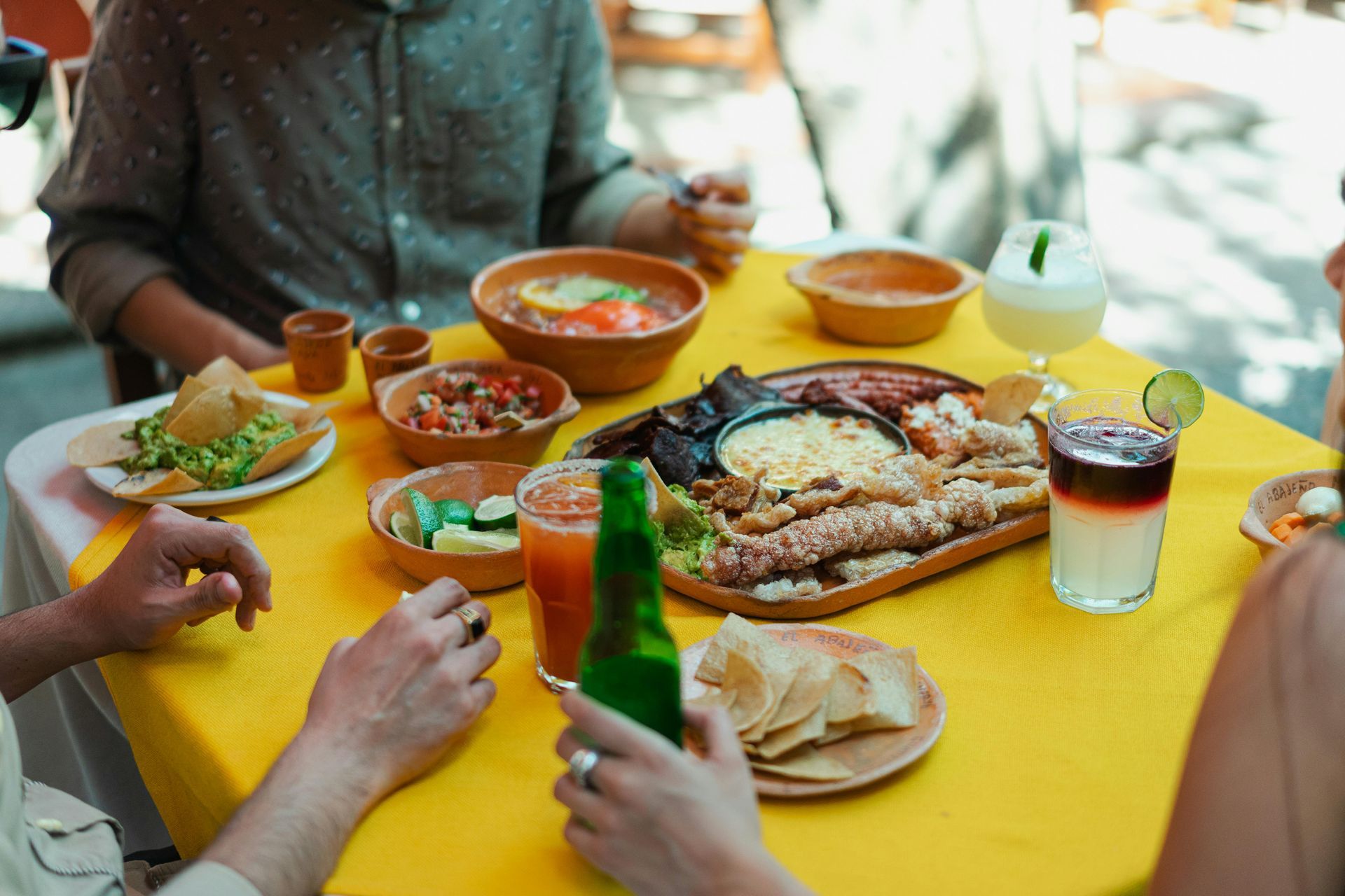 Table with Mexican food and drinks outdoors, yellow tablecloth. Hands reaching for food.