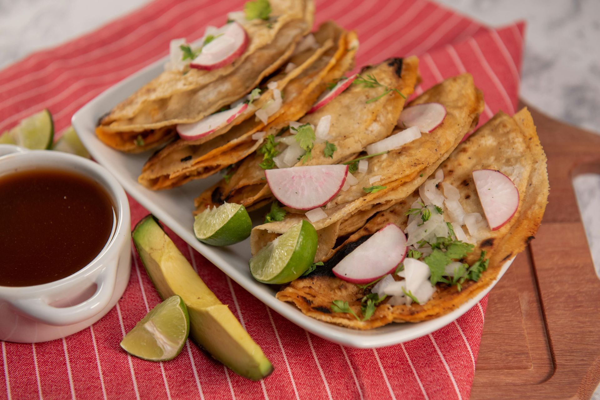 Four fried tacos topped with cilantro, onions, and radish slices on a white plate, served with lime, avocado, and salsa.
