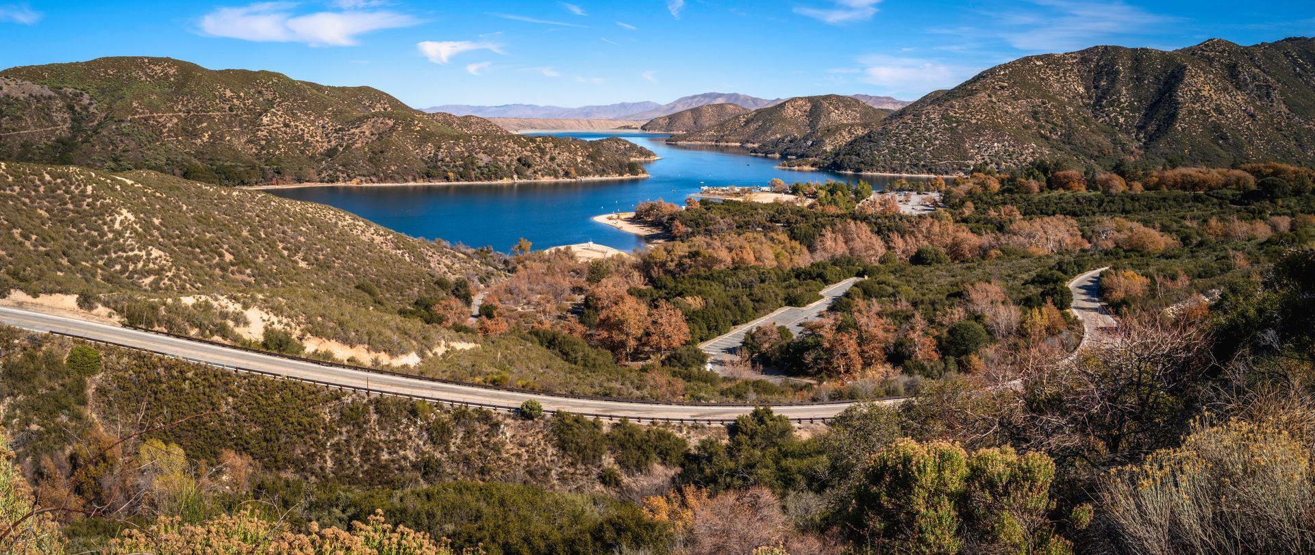 A winding road curves through a landscape of hills and dry trees toward a blue reservoir under a clear sky.