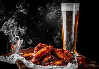A plate of steaming, saucy buffalo wings next to a tall glass of beer on a wooden board against a black background.