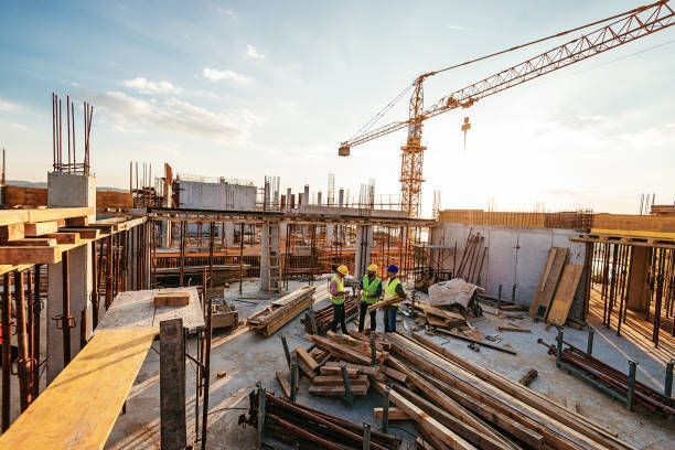 Three construction workers in hard hats and safety vests stand on a concrete floor at a sunny building site with a crane.