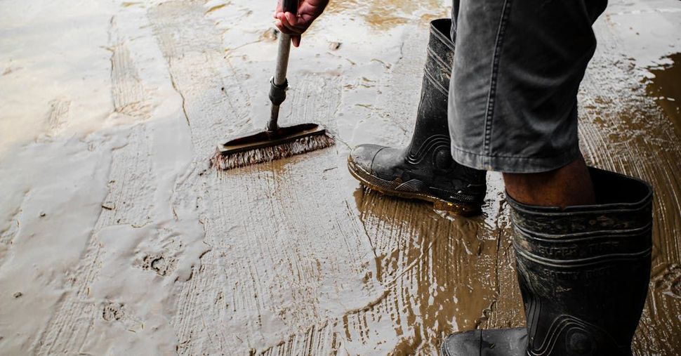 A person wearing rubber boots is cleaning a muddy floor with a broom.