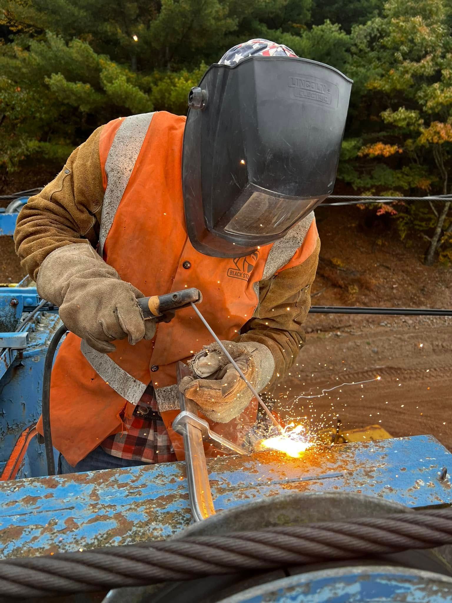 Welder in safety gear welding metal outdoors; sparks fly.