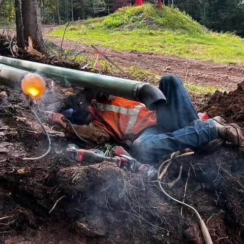Welder in an excavation, working on a pipe. Orange vest, safety gear; sparks and smoke.