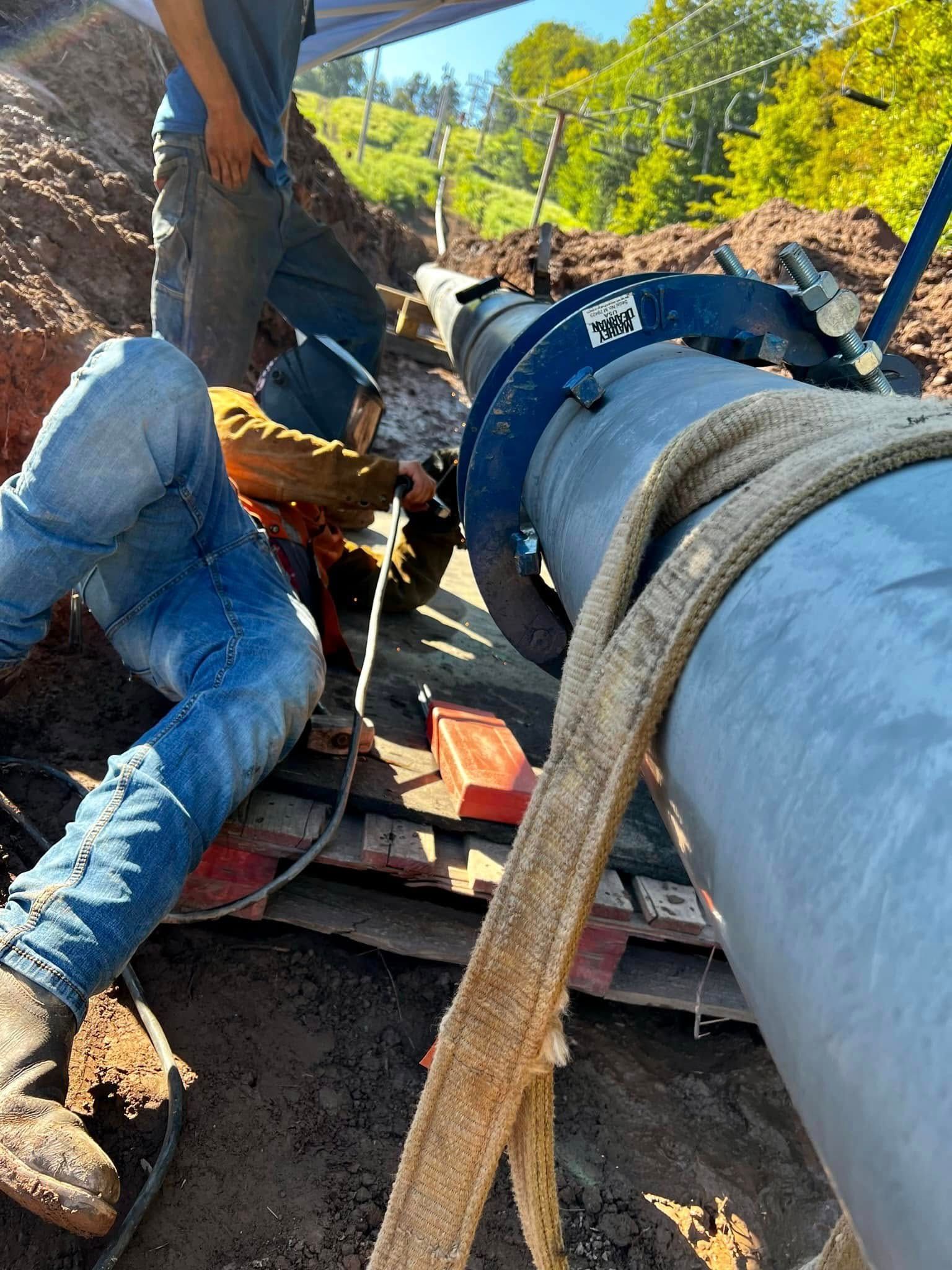 Workers welding a large pipe in a trench. One worker wears a welding mask.