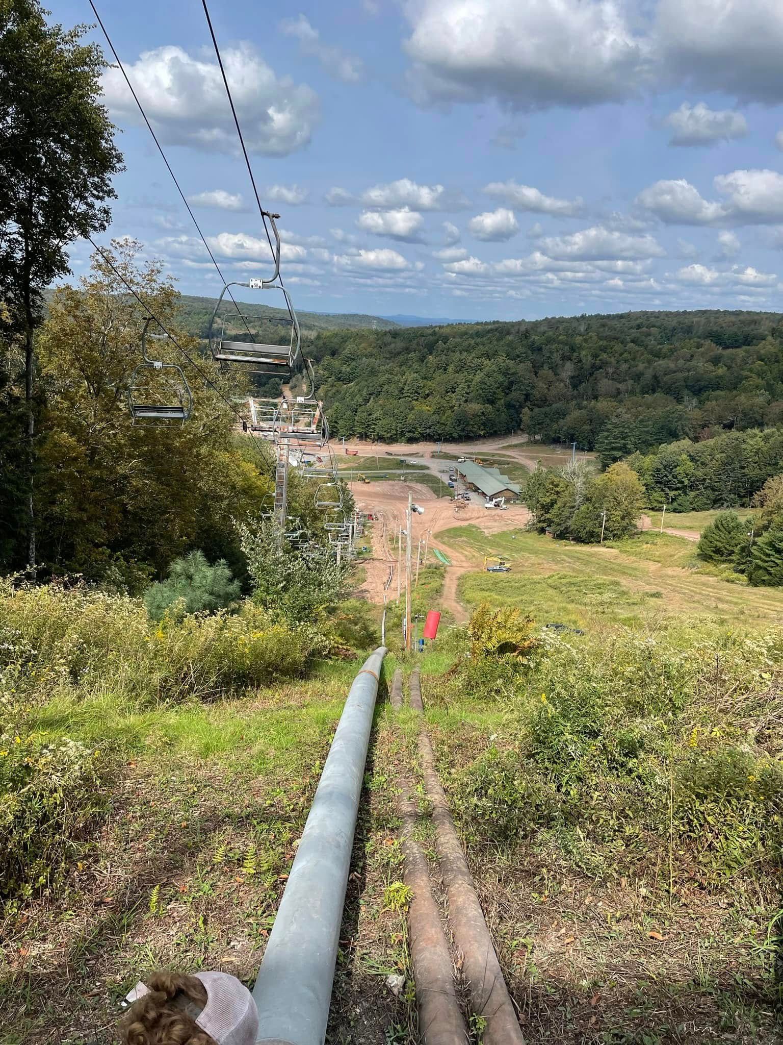 Ski lift over a grassy slope; buildings and trees in the distance under a partly cloudy sky.
