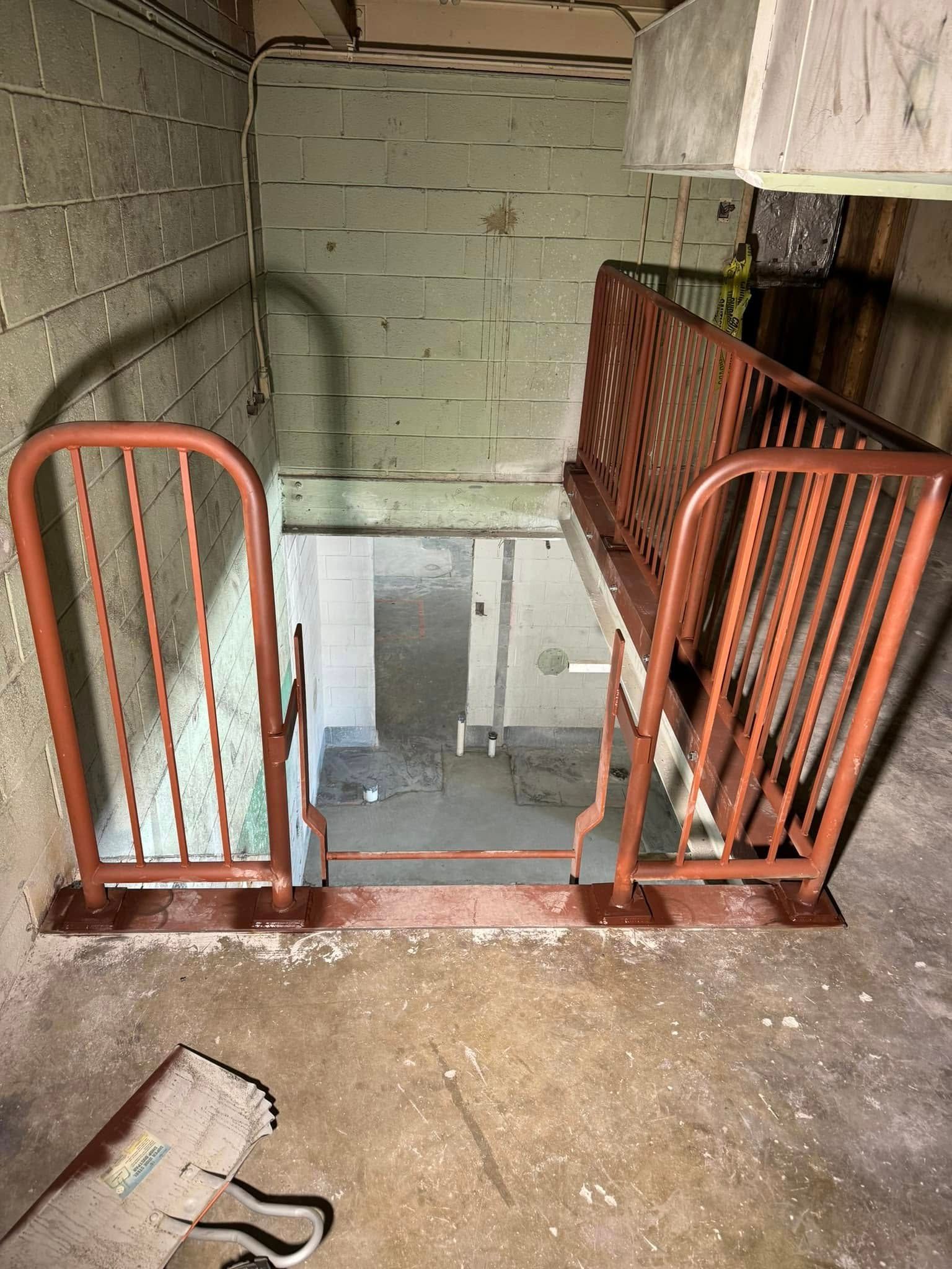 View down stairs with orange metal railings, leading to a dimly lit basement.