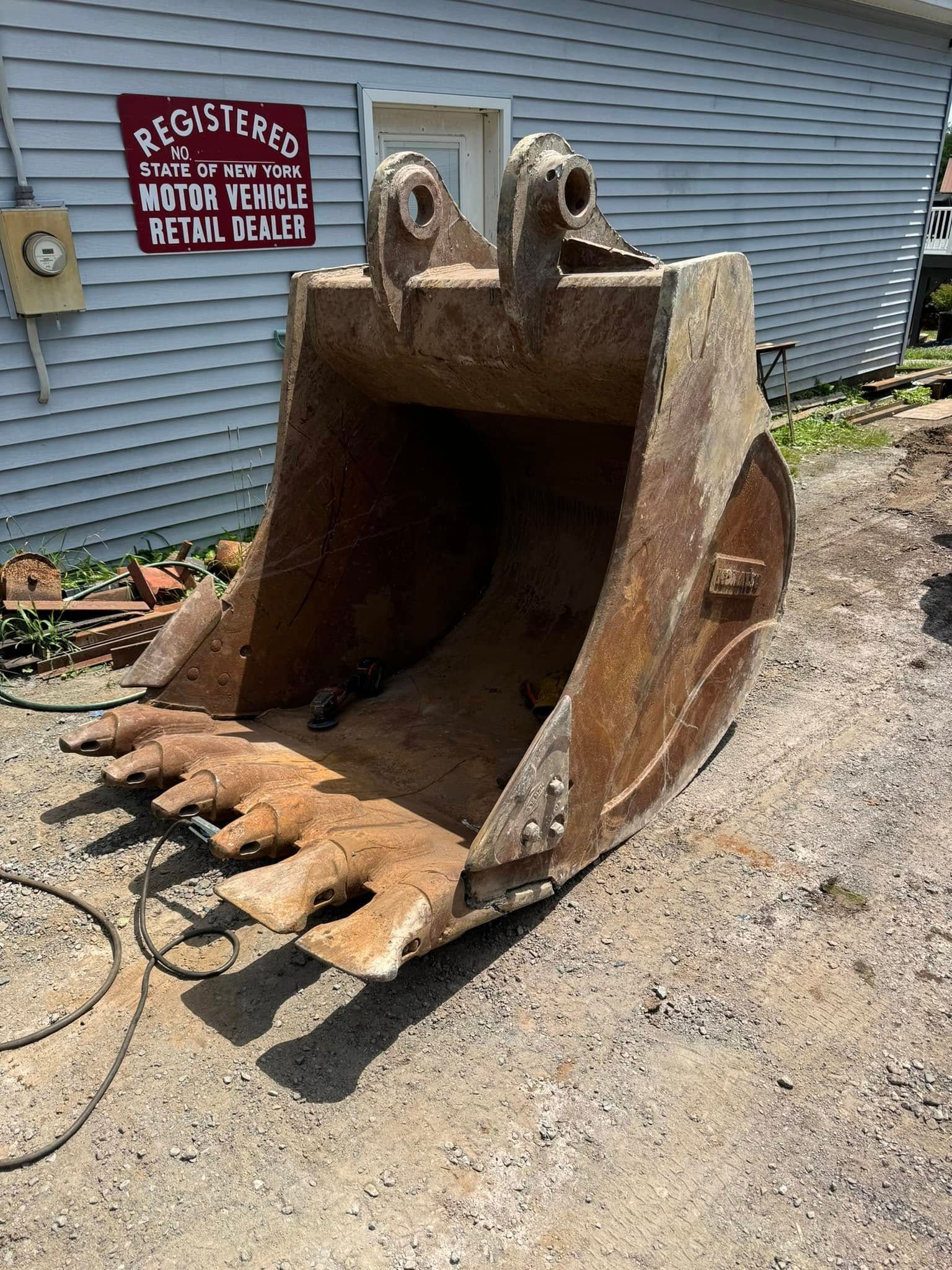 A large, rusty excavator bucket on a gravel surface, near a light blue building with a red sign.