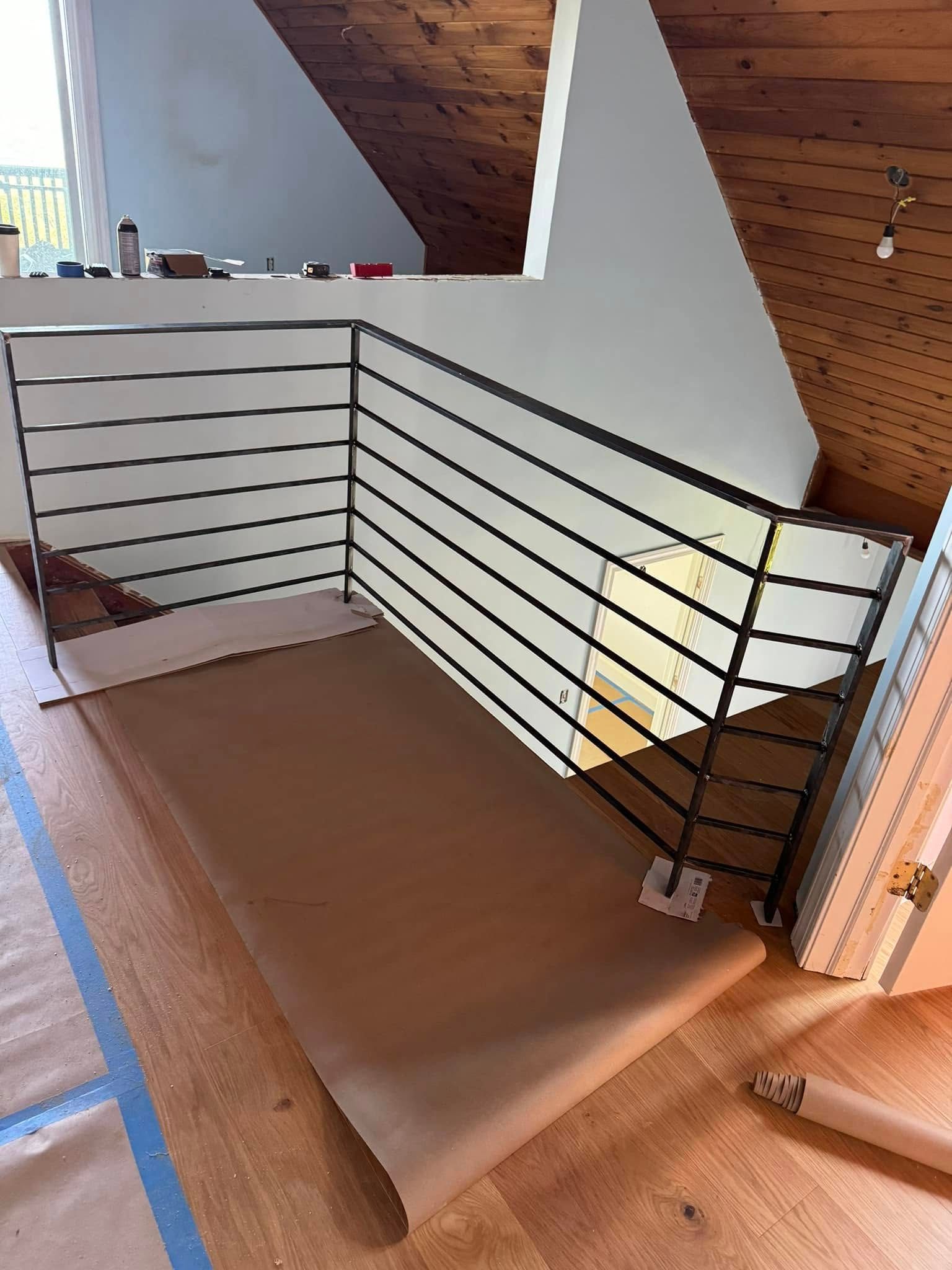 Metal railing installed on a wooden floor, protecting a stairwell. Brown paper covers floor near railing.