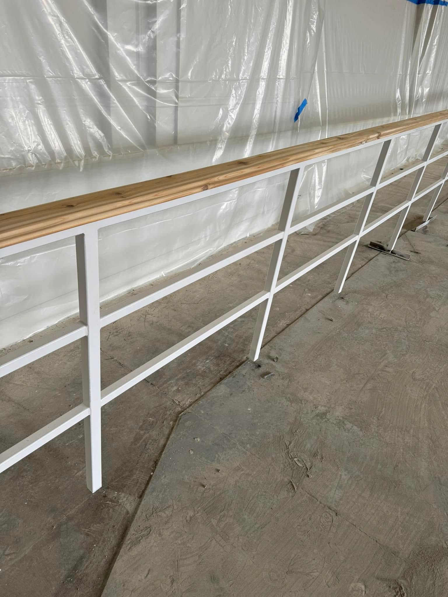 White metal railing with wooden top, against a white wall and concrete floor.