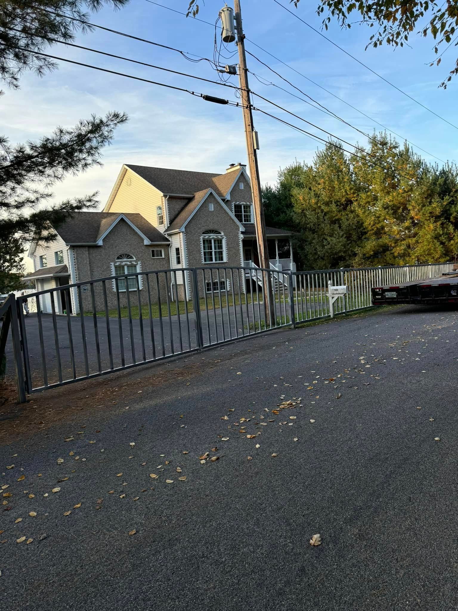 Gray stone house behind a wrought iron gate on a gravel driveway, with power lines and trees in the background.
