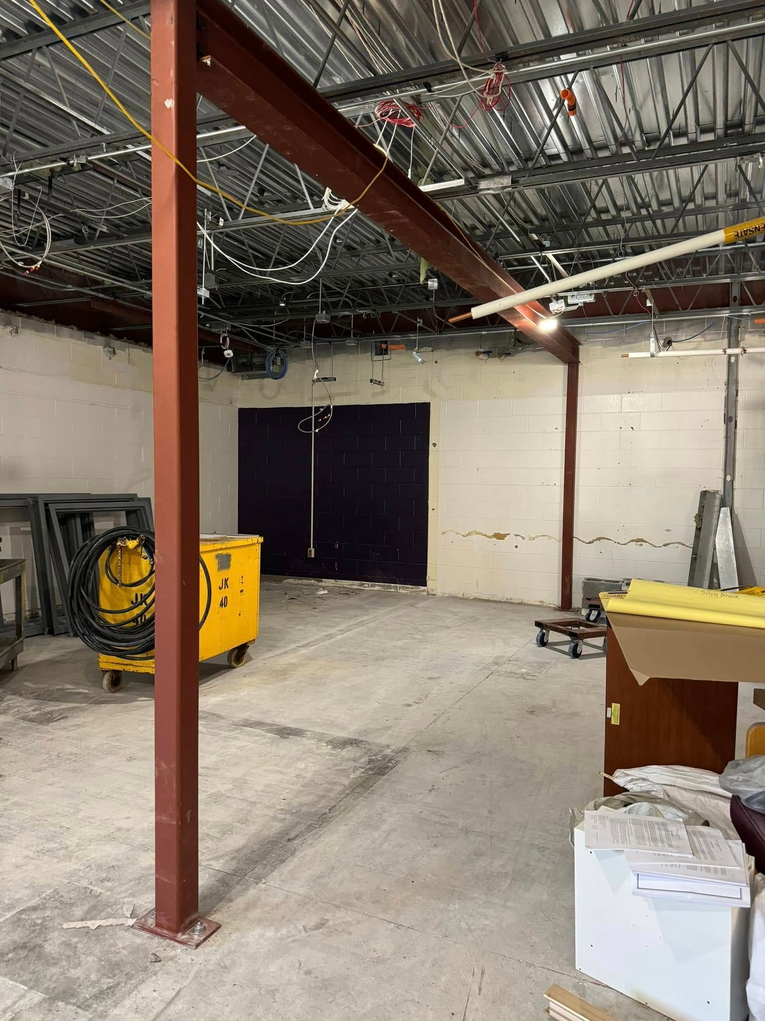 Interior of a construction site with a red metal support beam, yellow equipment, and concrete floor.