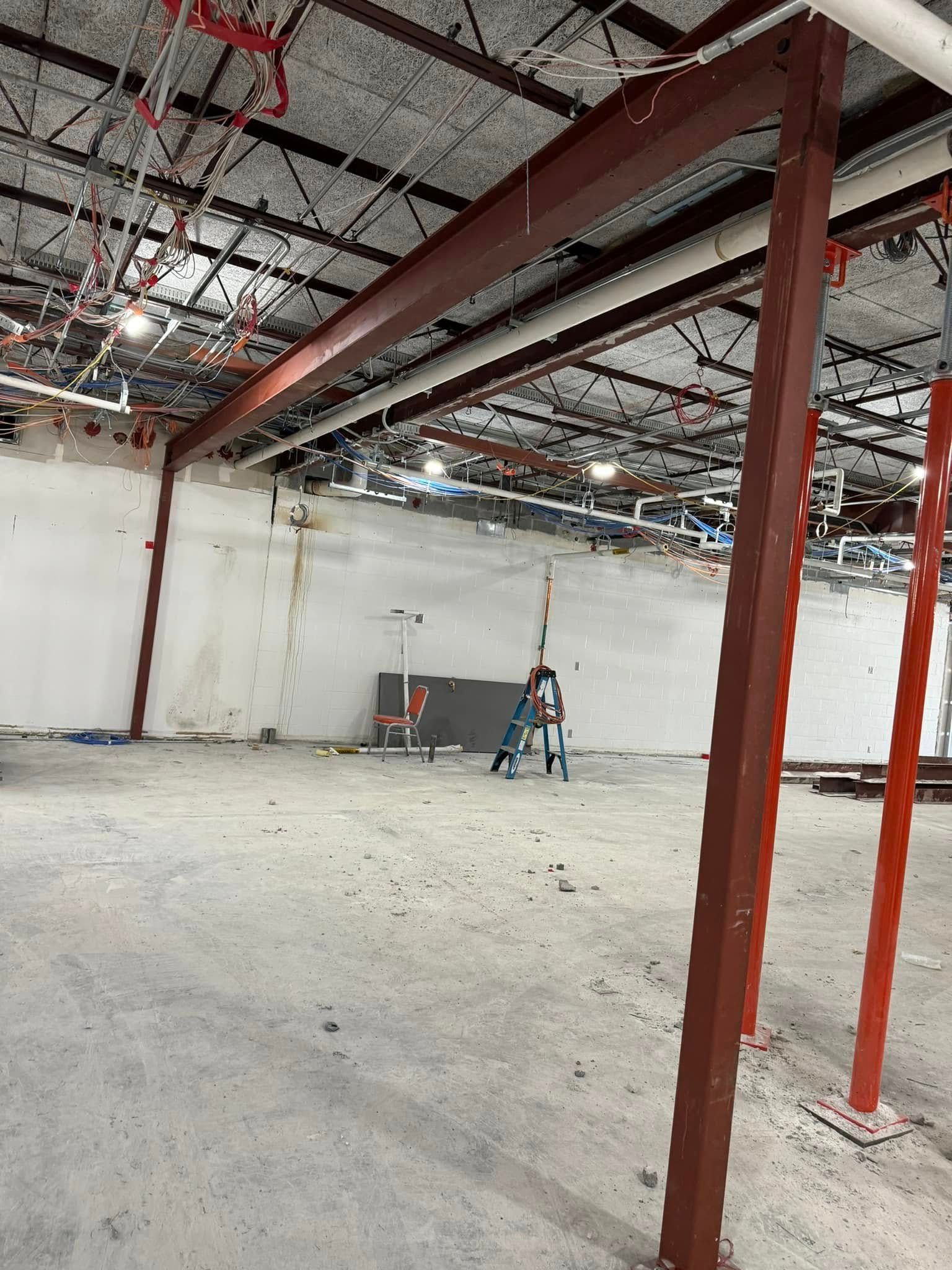 Construction site interior: red steel beams, bare concrete floor, exposed pipes, white walls, and bright overhead lighting.