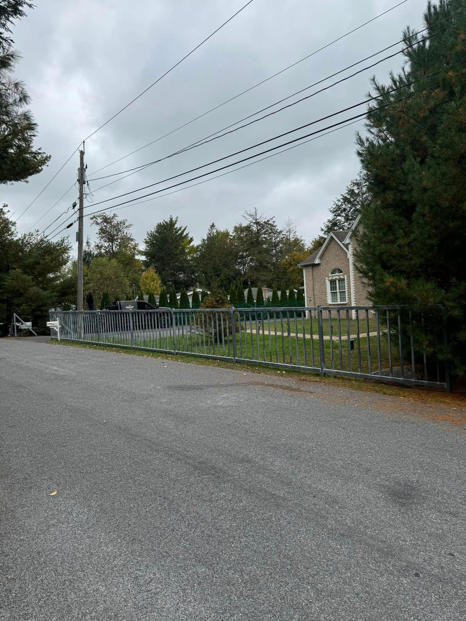 Gravel driveway leading to a house with a decorative fence and trees under a cloudy sky.