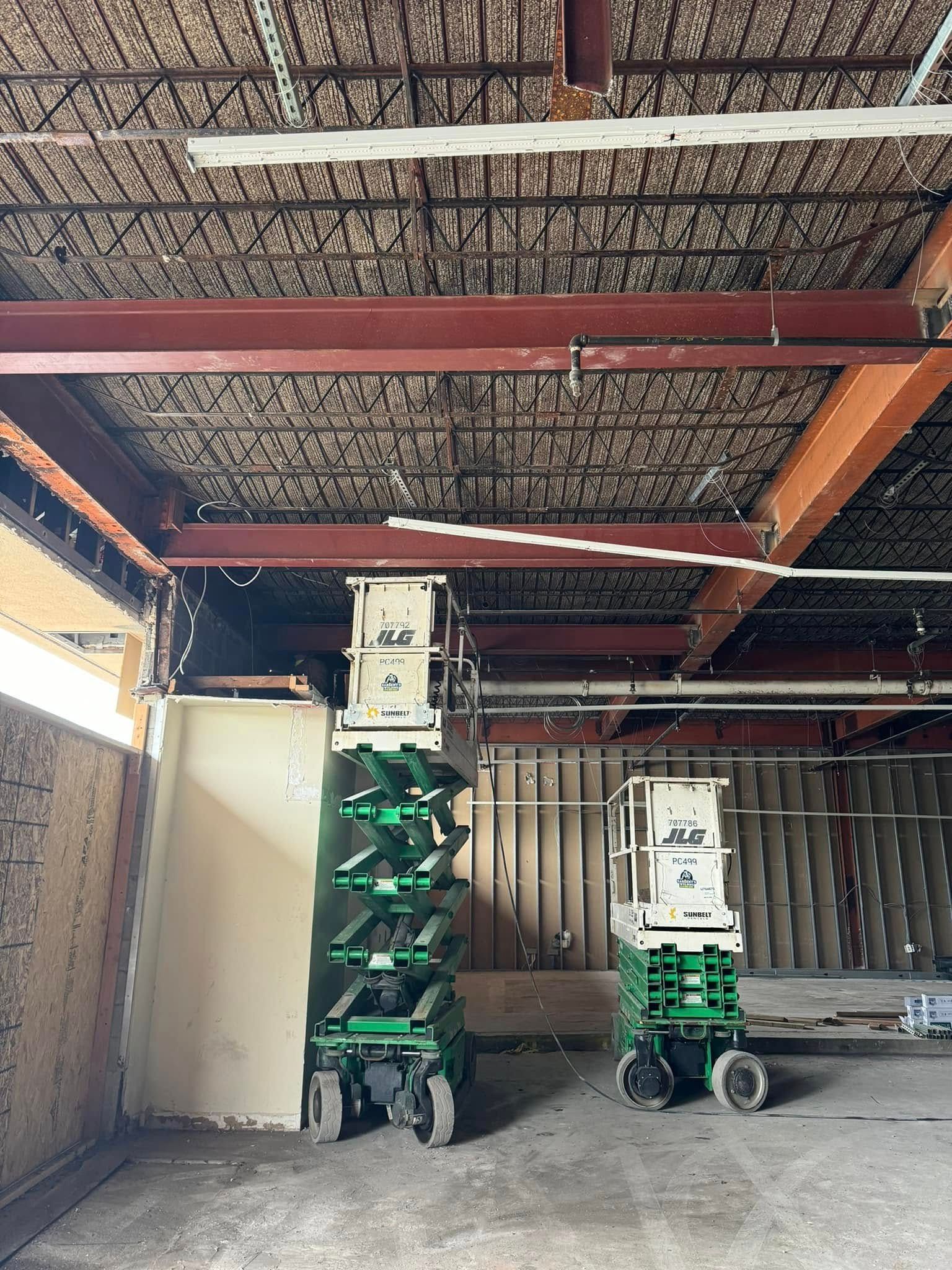 Two green scissor lifts under a ceiling with exposed beams, in a building during construction.