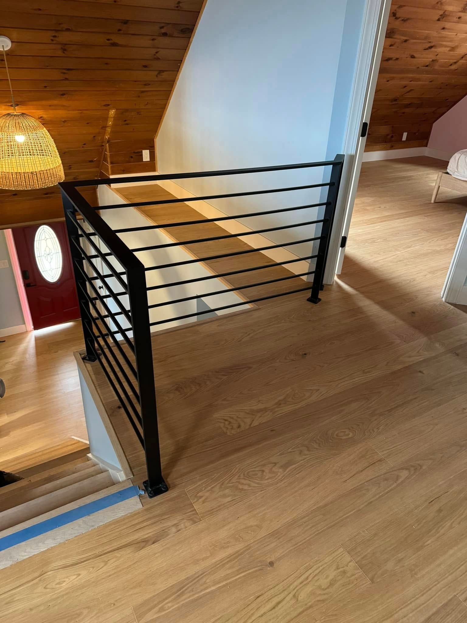 Black metal railing on a wooden floor landing, leading to a staircase. Soft lighting, open space.