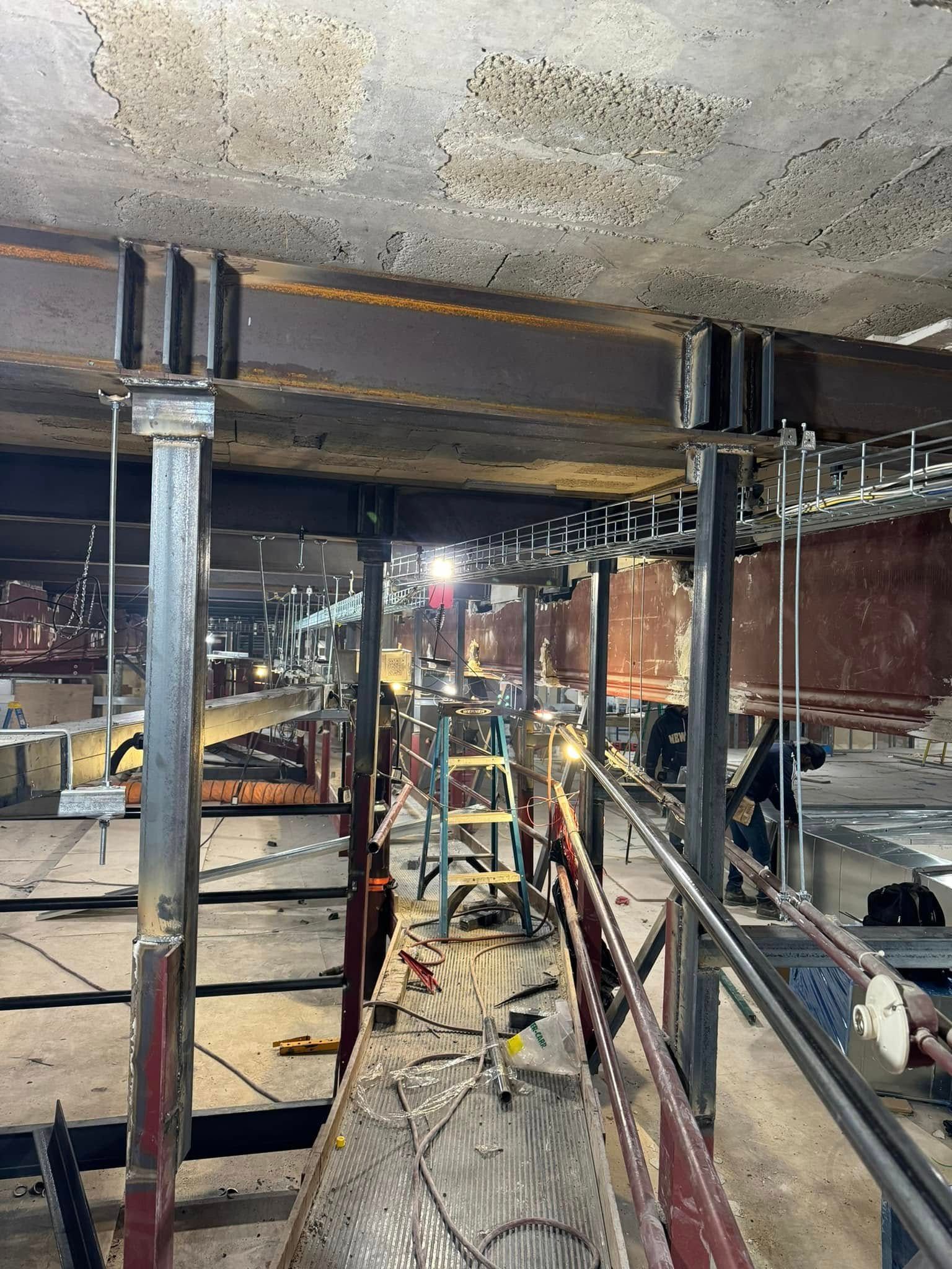 Construction site: Steel beams supporting a ceiling. Workers on scaffolding, cables and tools present.