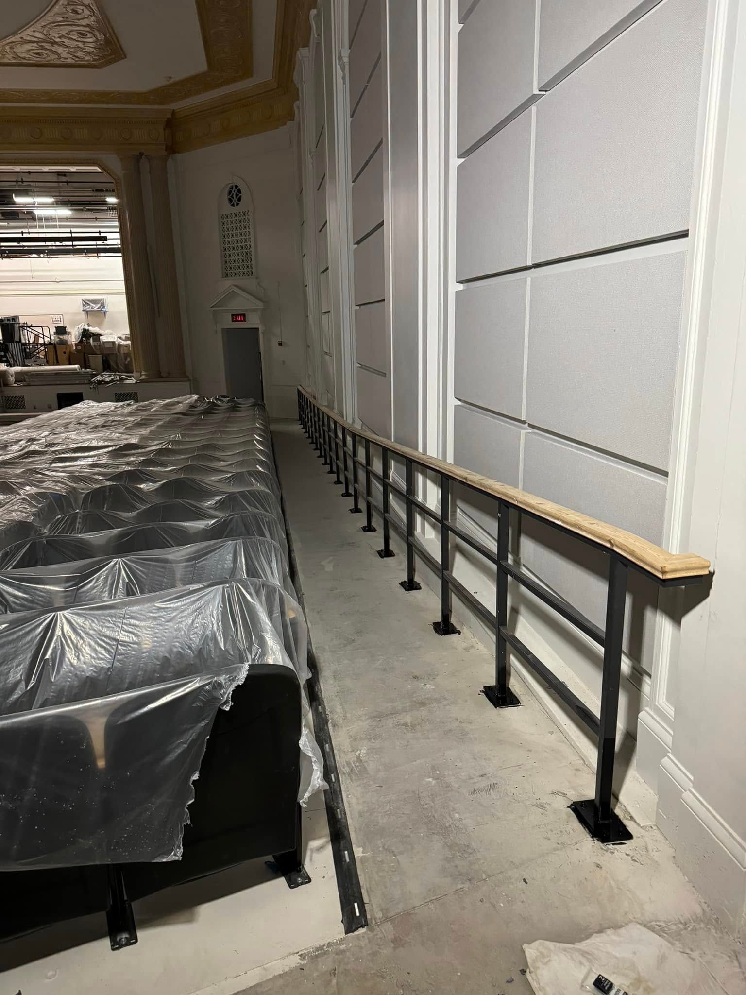 Theater interior with a newly installed handrail along a hallway. Black metal railing, wood top, and light gray walls.
