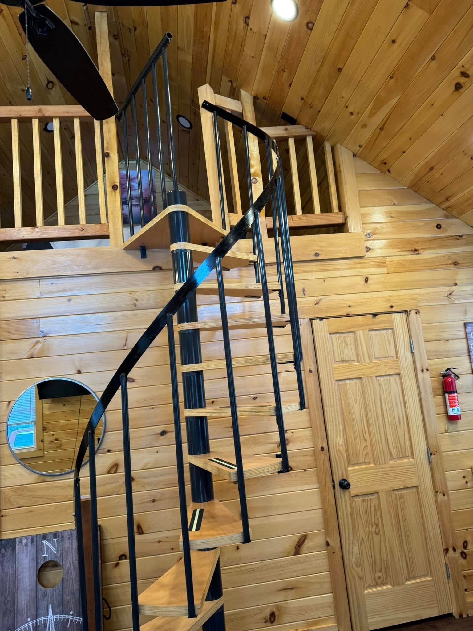 Spiral staircase in a wooden cabin leads to a loft with a wooden railing.