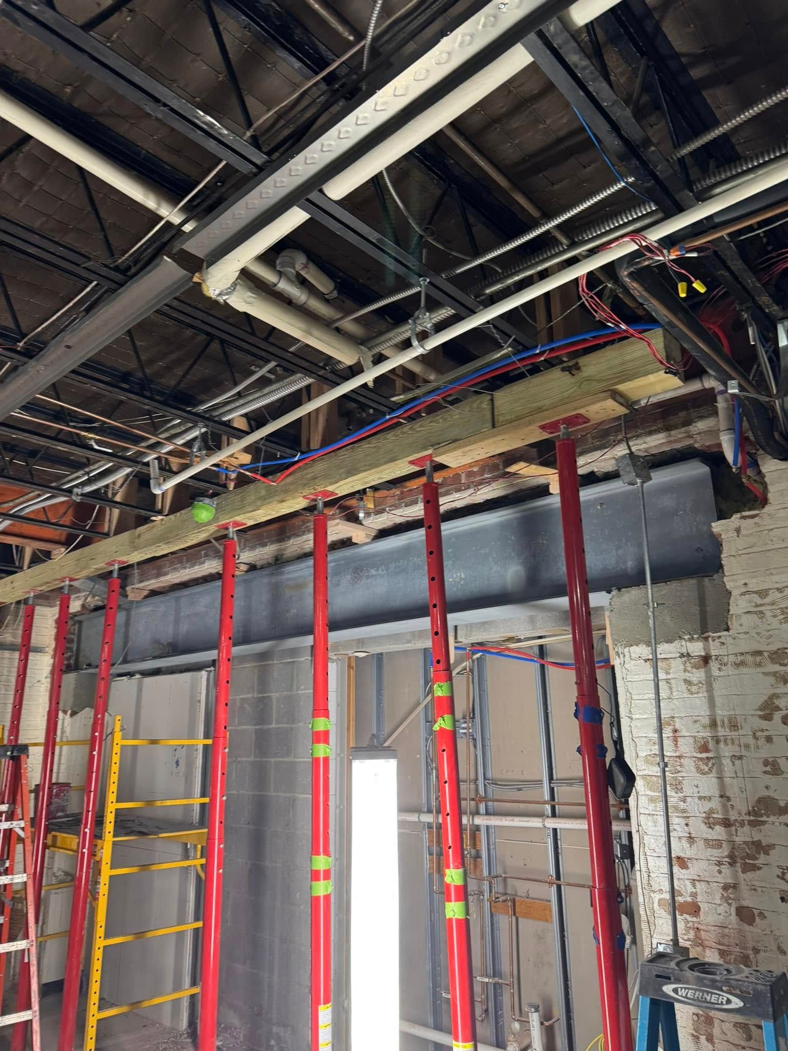 Construction site. Steel beam supported by red jacks. Exposed ceiling. Brick wall partially visible.