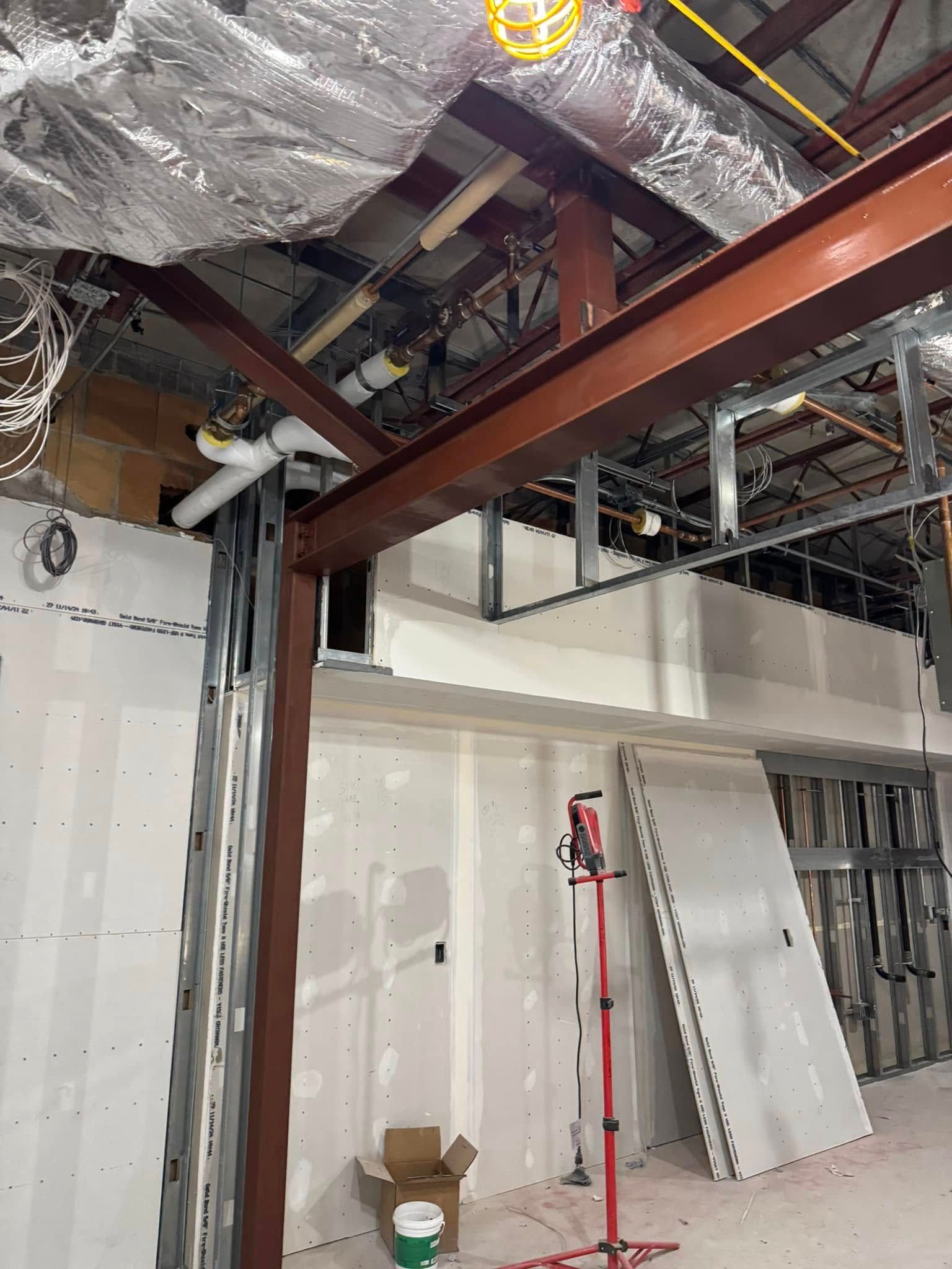 Construction site with exposed metal beams, drywall, and ductwork. Red steel supports and a work light are visible.