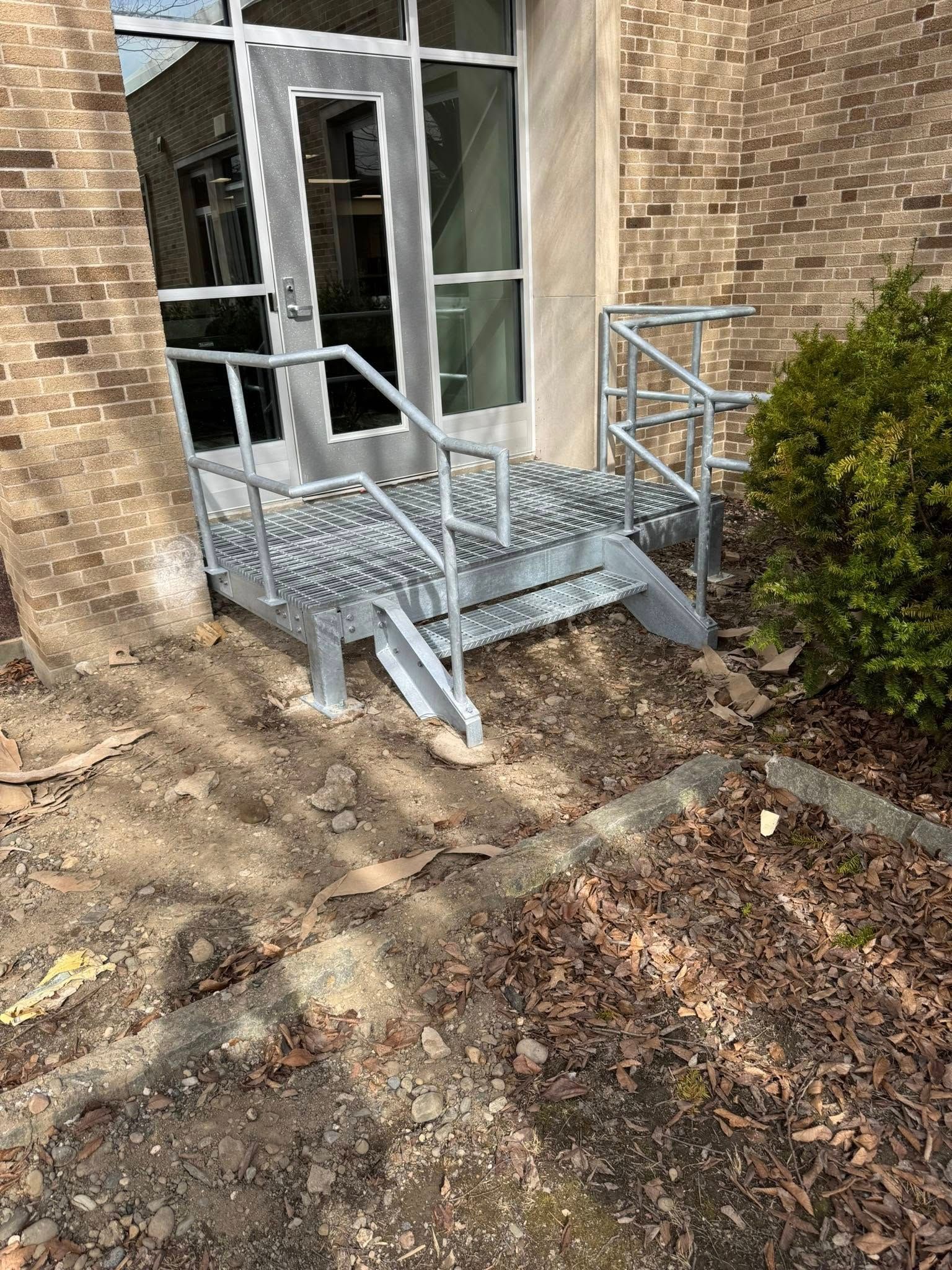 Metal ramp and steps leading up to a building entrance with glass door. Brown brick and dirt ground.