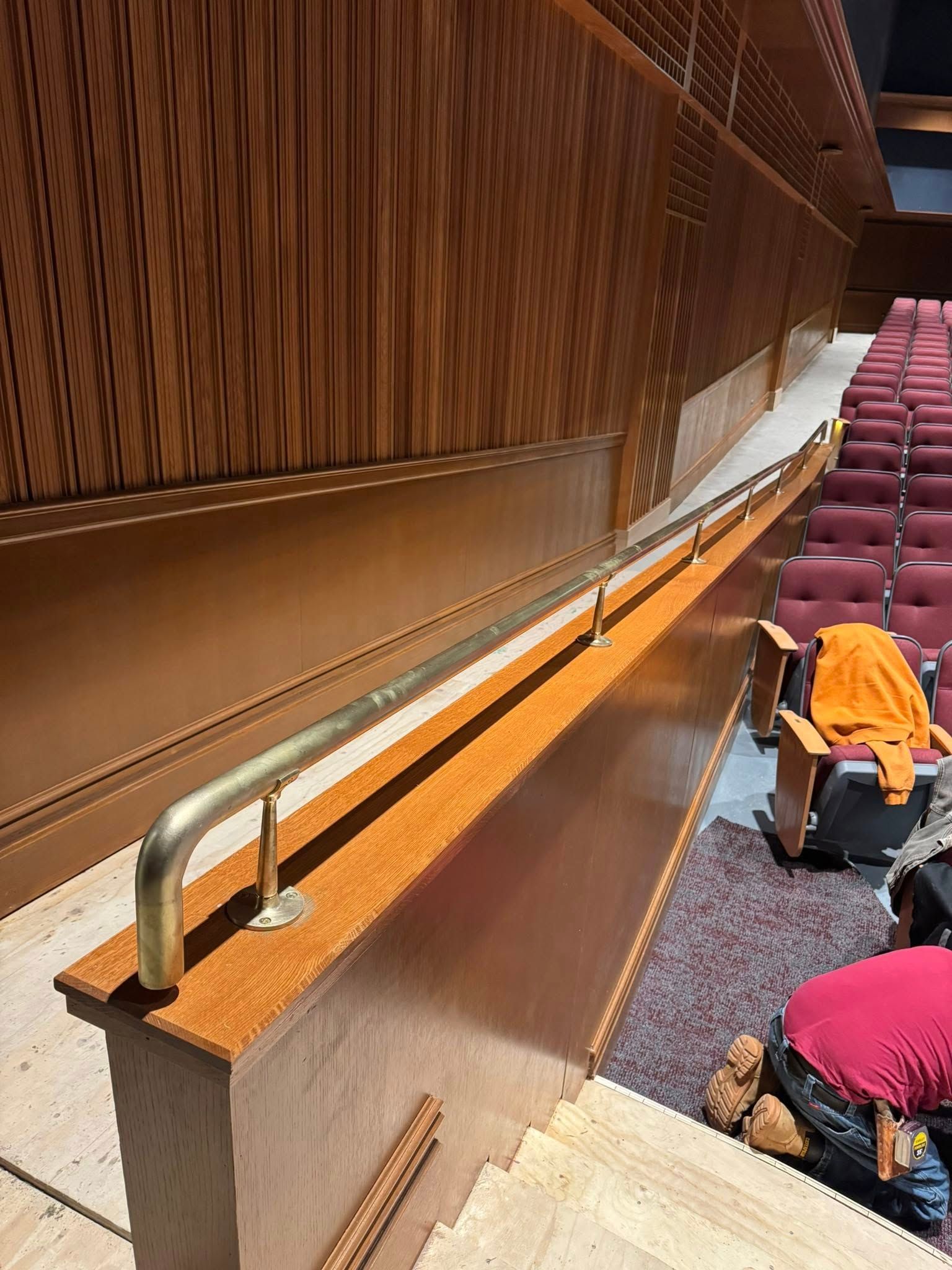 A handrail in an auditorium, with seating and wooden paneling. A person kneels below.
