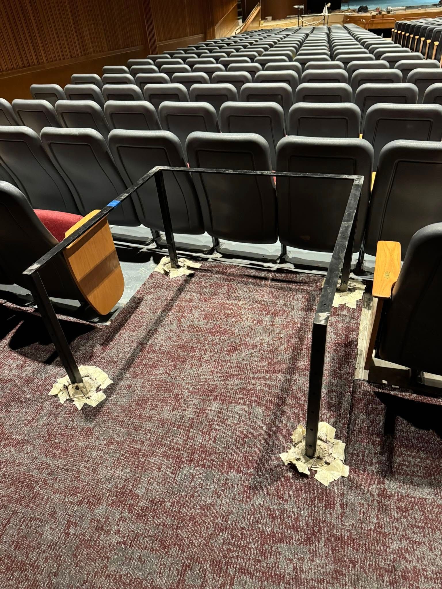 An empty auditorium with a makeshift metal handrail taped to the carpet in front of some seats.
