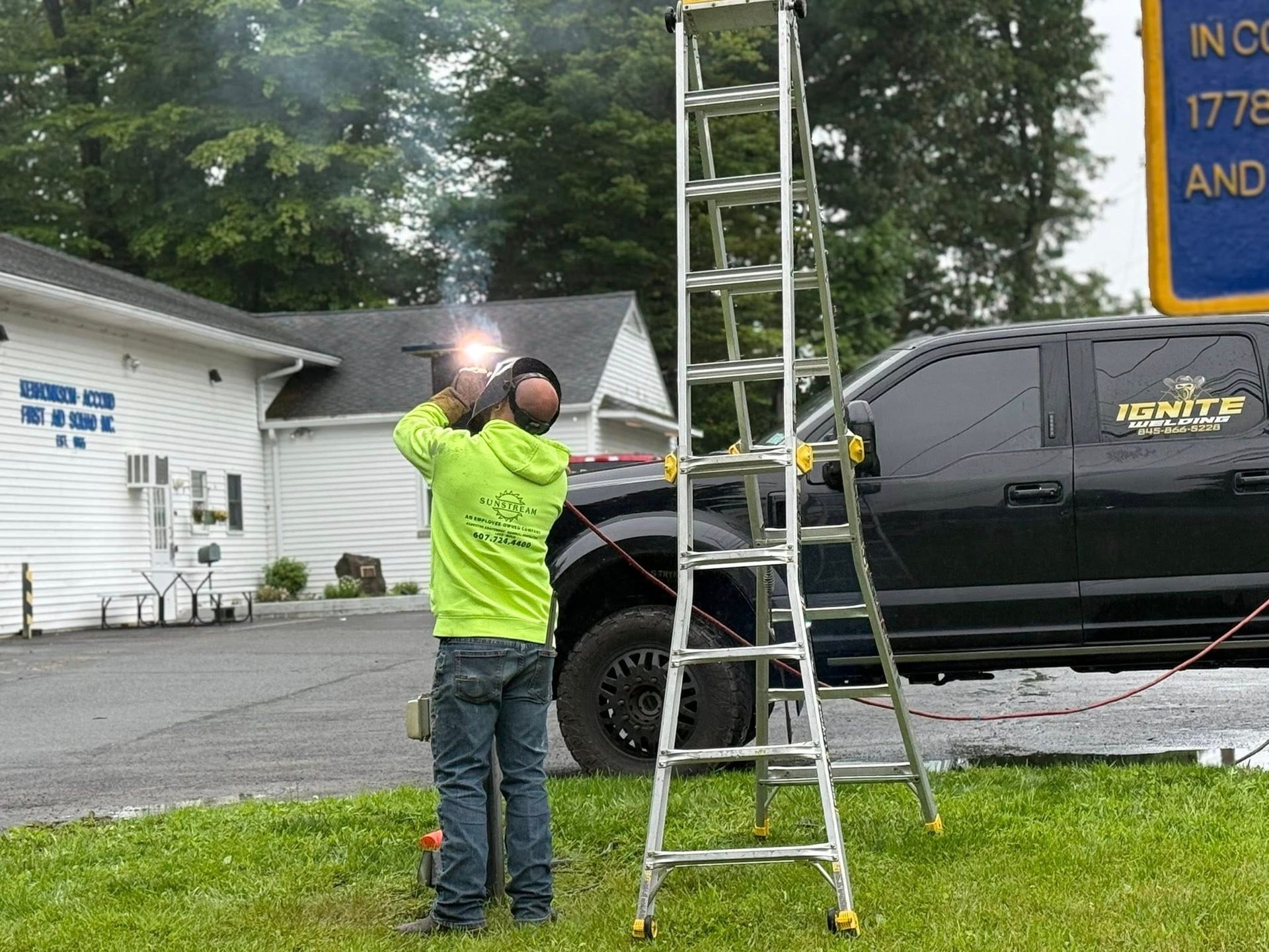 Man welding on a ladder outside a building; wearing a neon green hoodie. Black truck and sign visible.