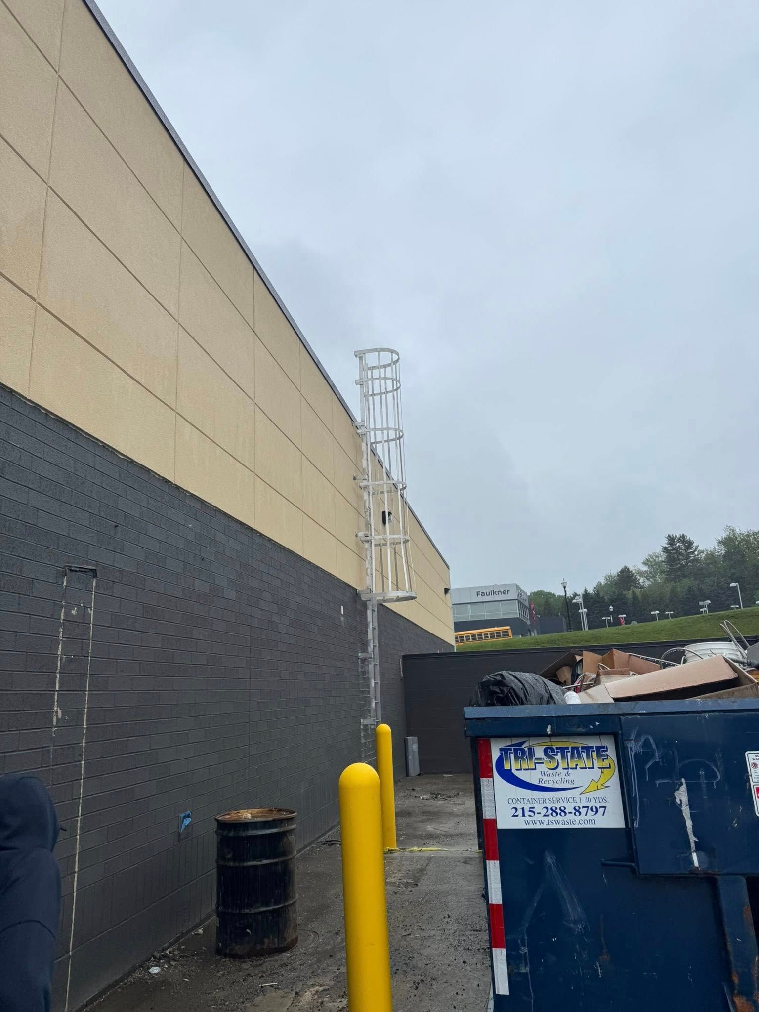 Exterior view of a building with a ladder. A dumpster and yellow posts are in front. Overcast sky.