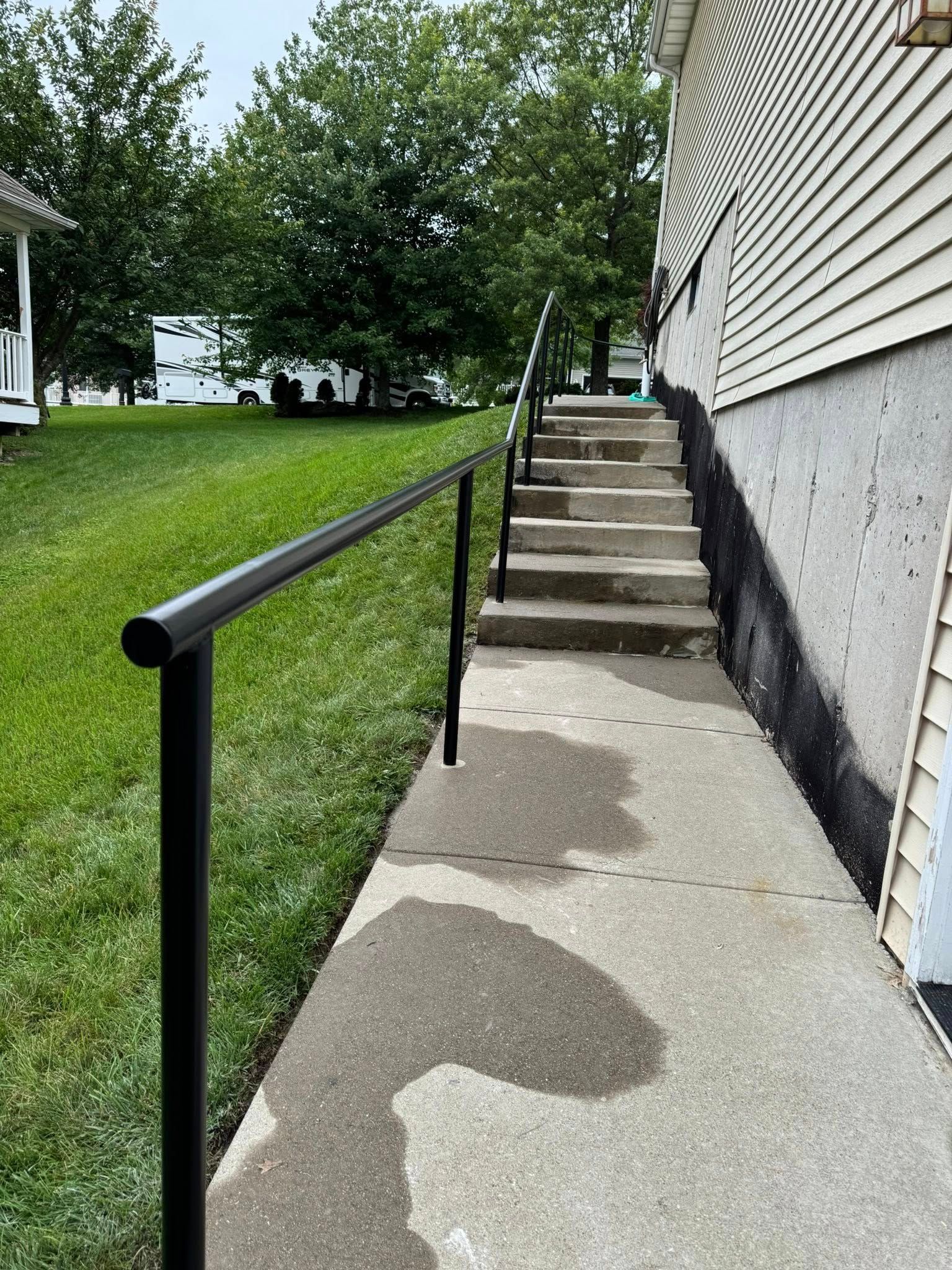 Black handrails on concrete steps leading to a building. Green grass and trees in the background.