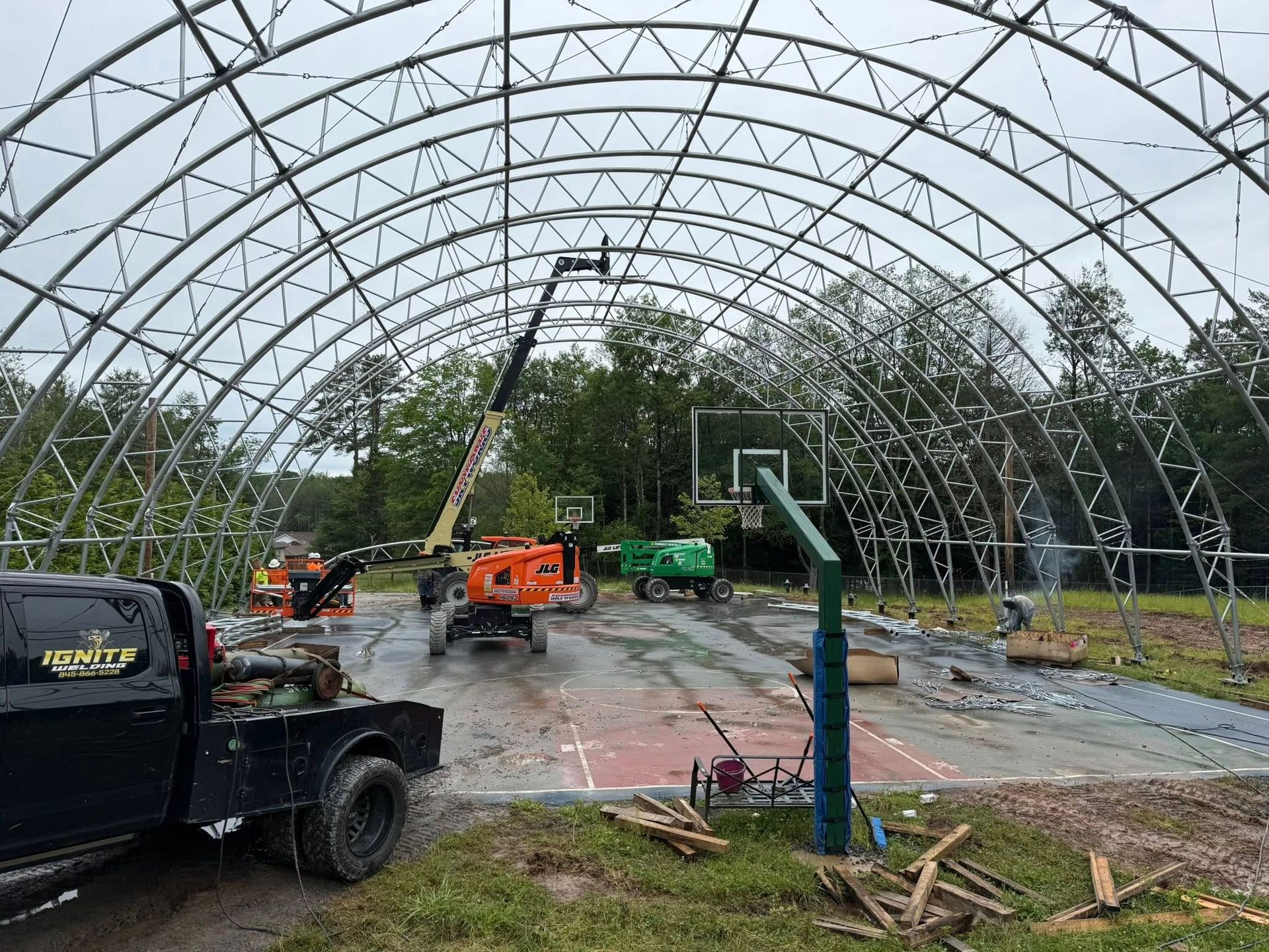 Construction of an arched steel building over a basketball court; several workers and machinery are present.