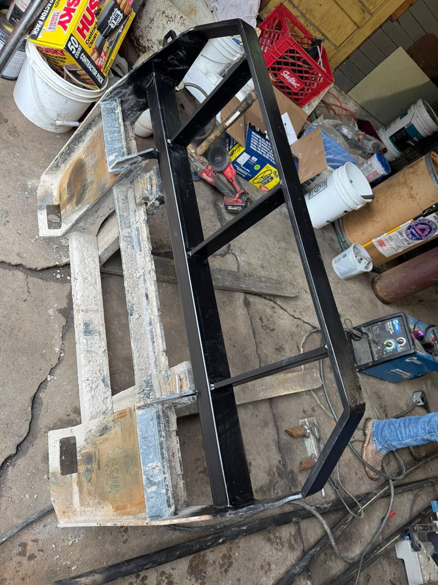 Metal frame welded to an old, rectangular structure; black frame, concrete floor, cluttered shop background.