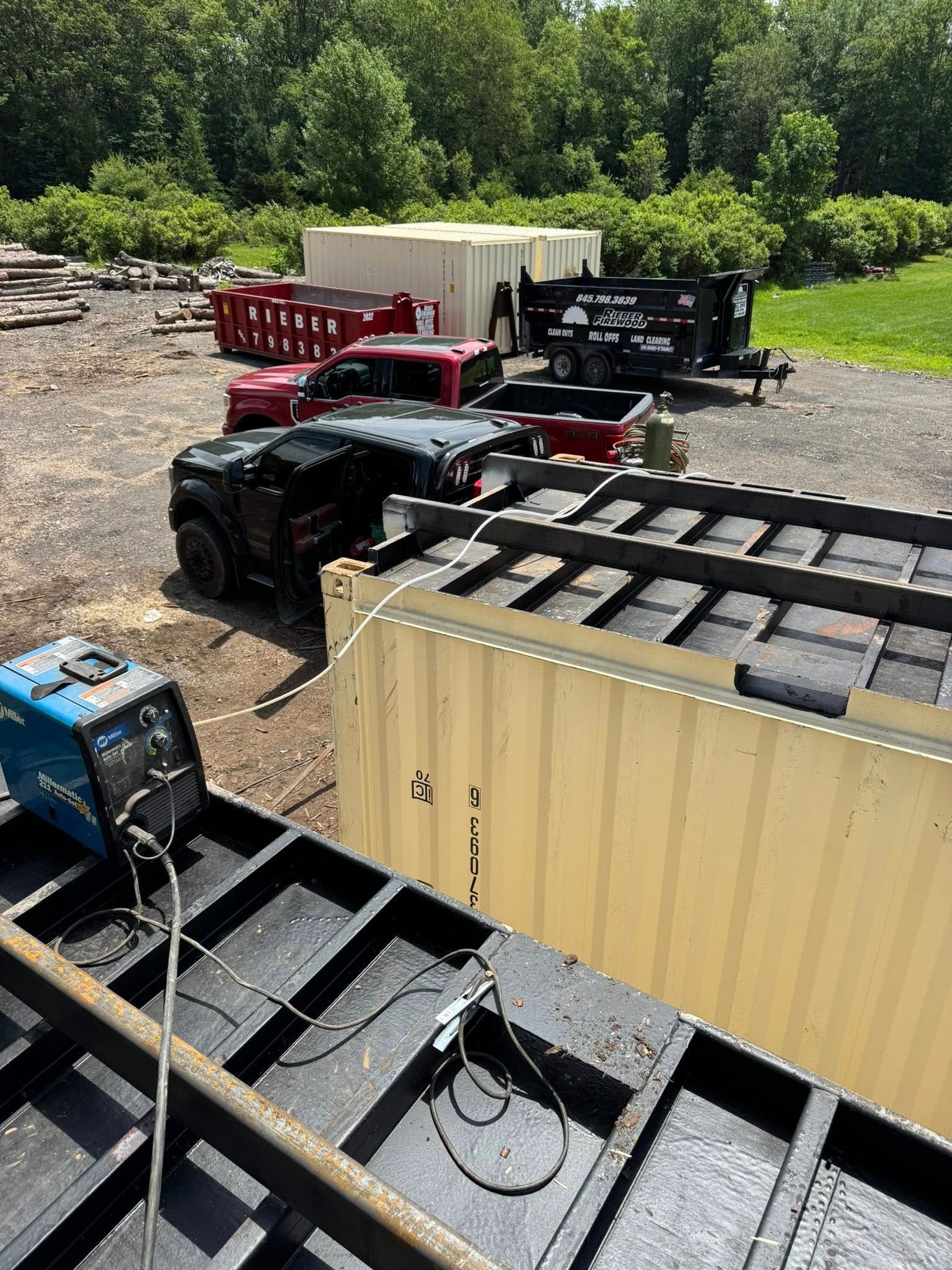 A blue welding machine on black metal frame. Vehicles and construction containers in a sunny outdoor setting.