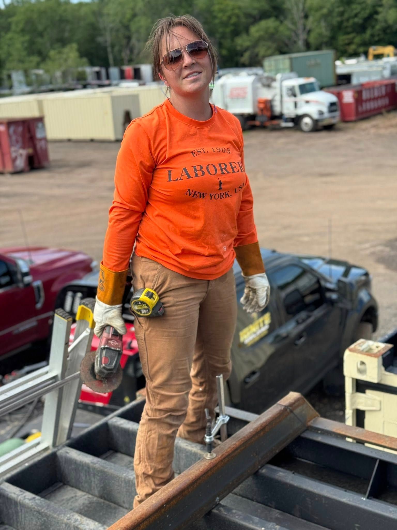 Woman in work clothes, holding a grinder, stands on a rooftop platform in an outdoor industrial setting.