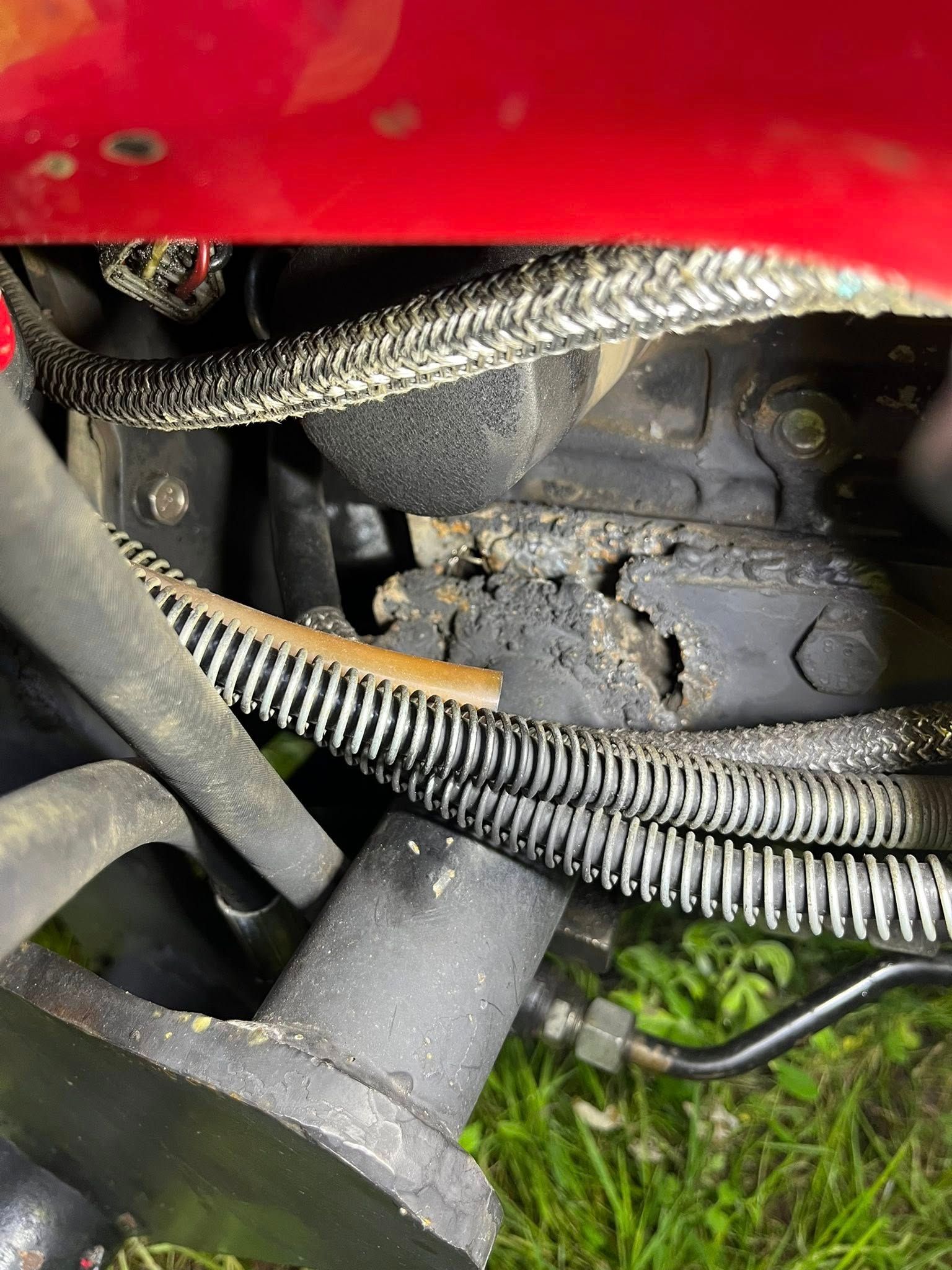 Red car engine bay with damaged black component; hoses and wiring visible.