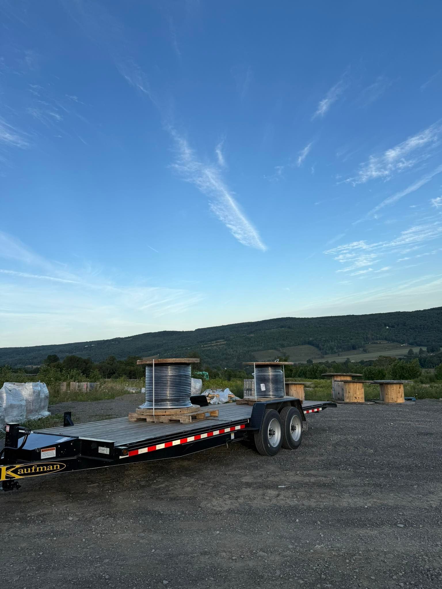 Flatbed trailer with spools of cable against a blue sky with wispy clouds and a green hillside.