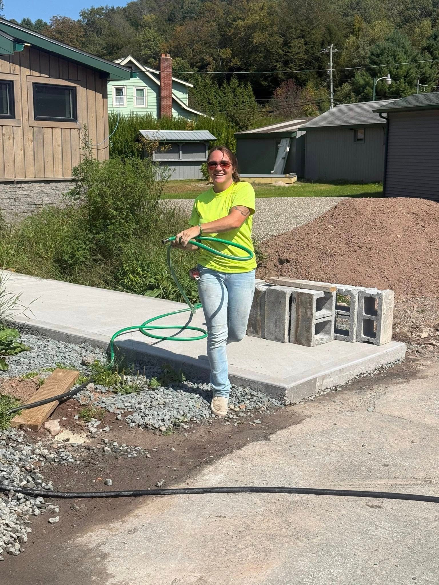 Woman in yellow shirt and jeans holds green hose, standing on concrete, with cinder blocks nearby.