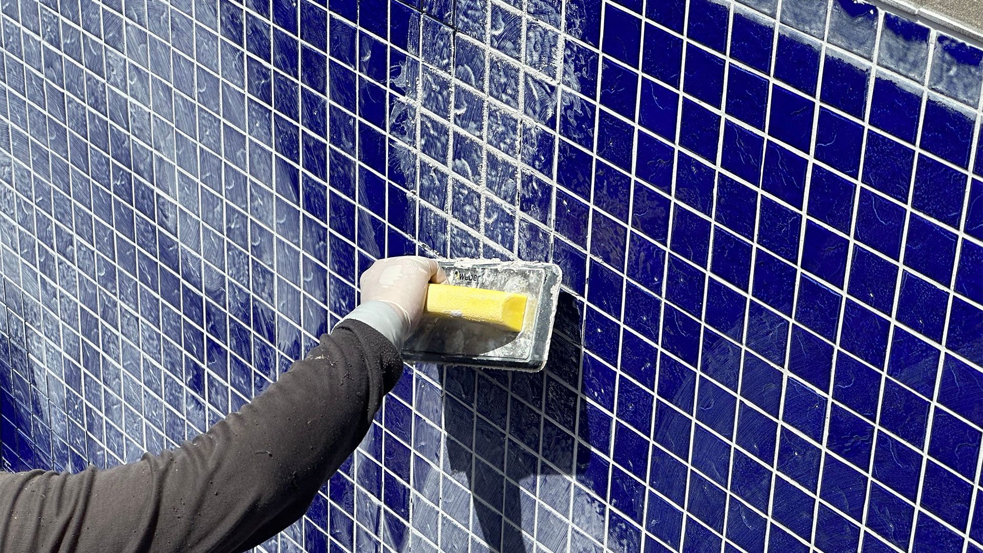 Person grouting blue diamond-patterned tiles on a wall, using a yellow sponge.