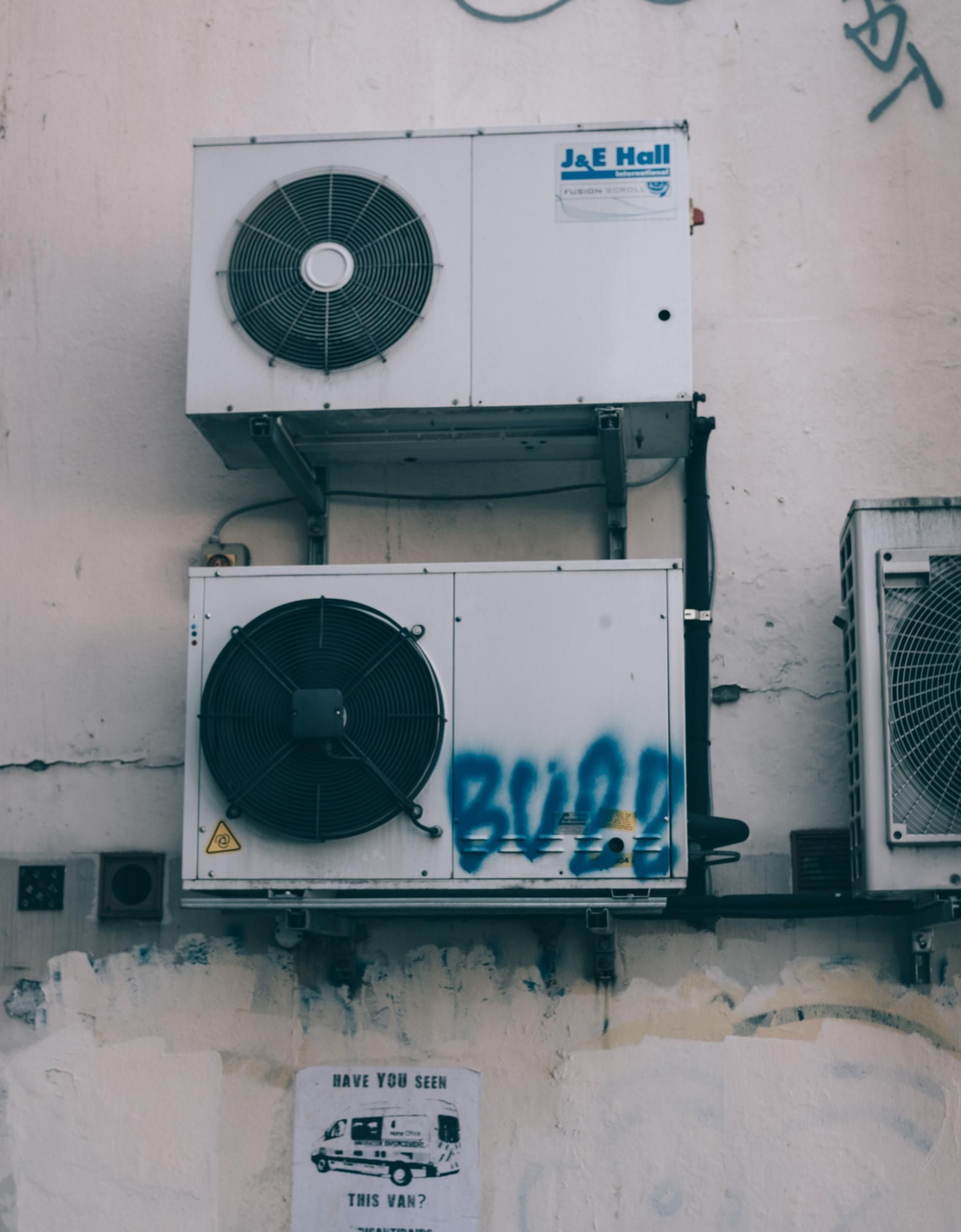 Two white air conditioning units stacked on a weathered wall, with graffiti.