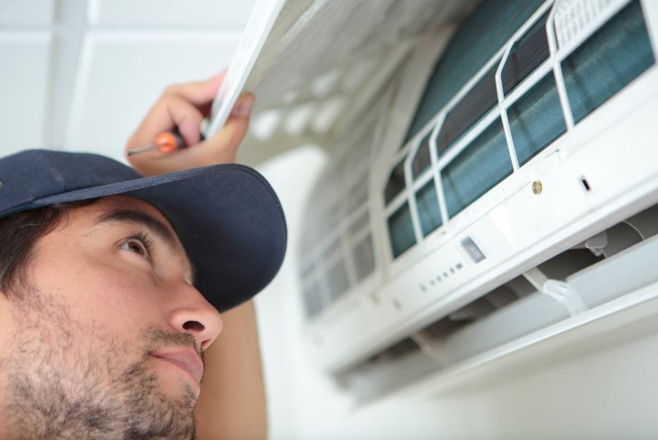 Man in cap repairing air conditioner with a screwdriver, looking upward.