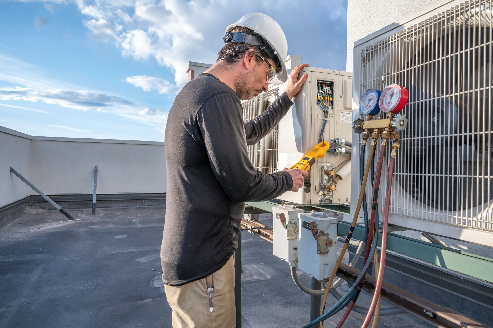 HVAC technician working on outdoor AC unit on a rooftop. He wears a hard hat and uses tools.