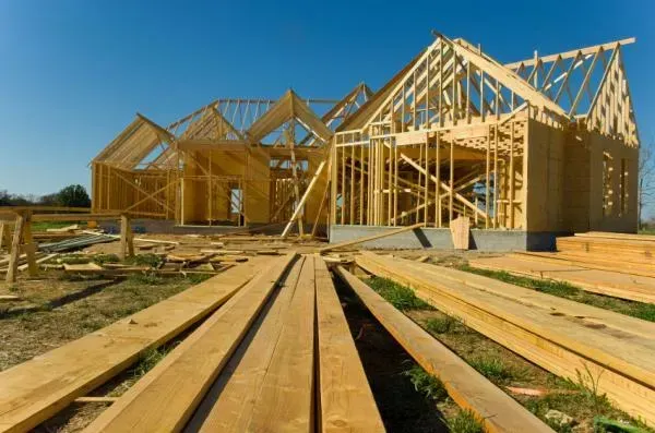 Wooden frame of a house under construction on a grassy lot, with stacks of lumber in foreground.