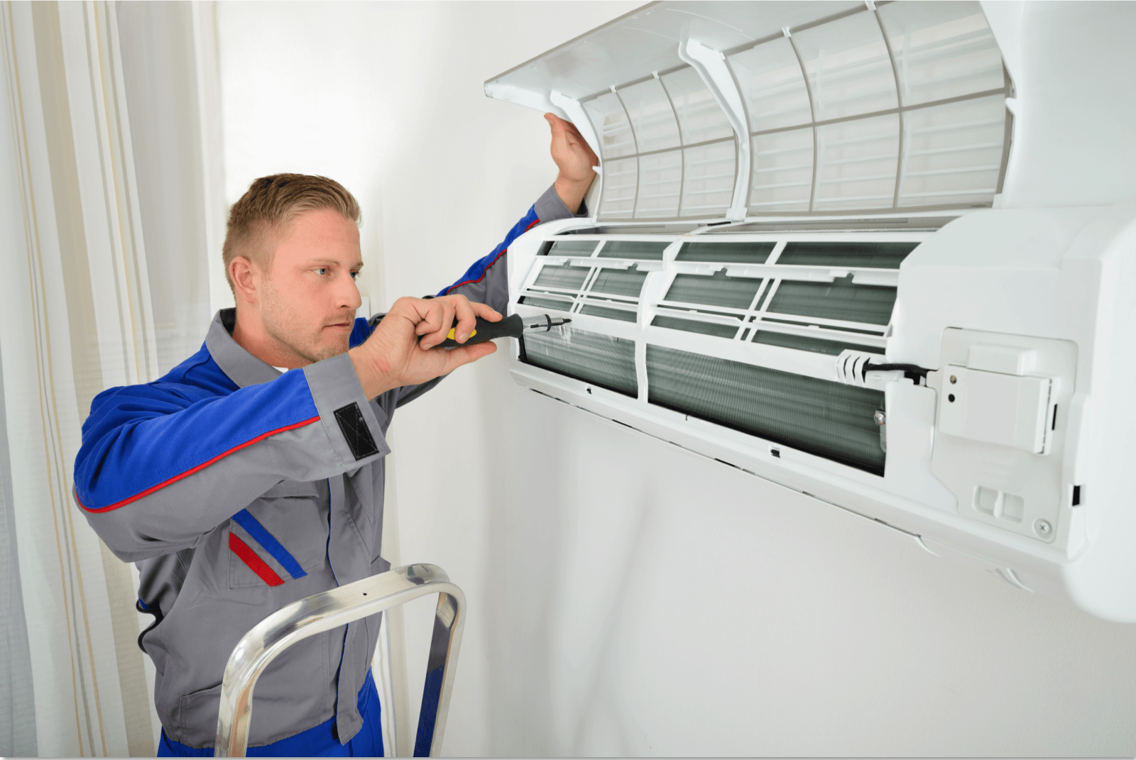 Man in blue jumpsuit repairs an air conditioner unit on a wall, using a screwdriver.