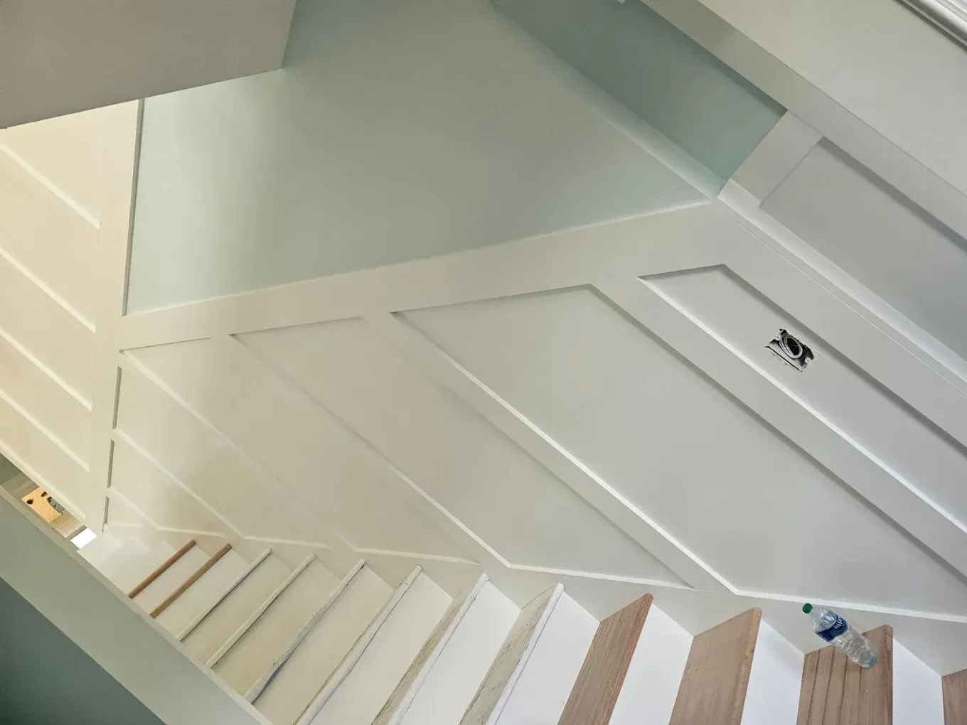 Staircase with white paneled walls and light blue walls above, viewed from above. Wooden steps lead downwards.