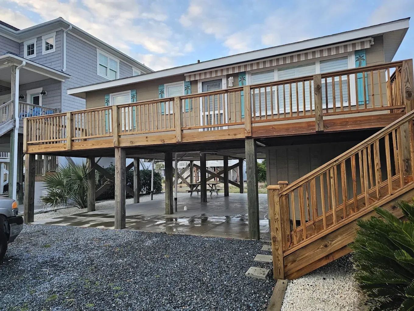 Beach house with wooden deck on stilts. Exterior view shows a staircase and gravel yard.